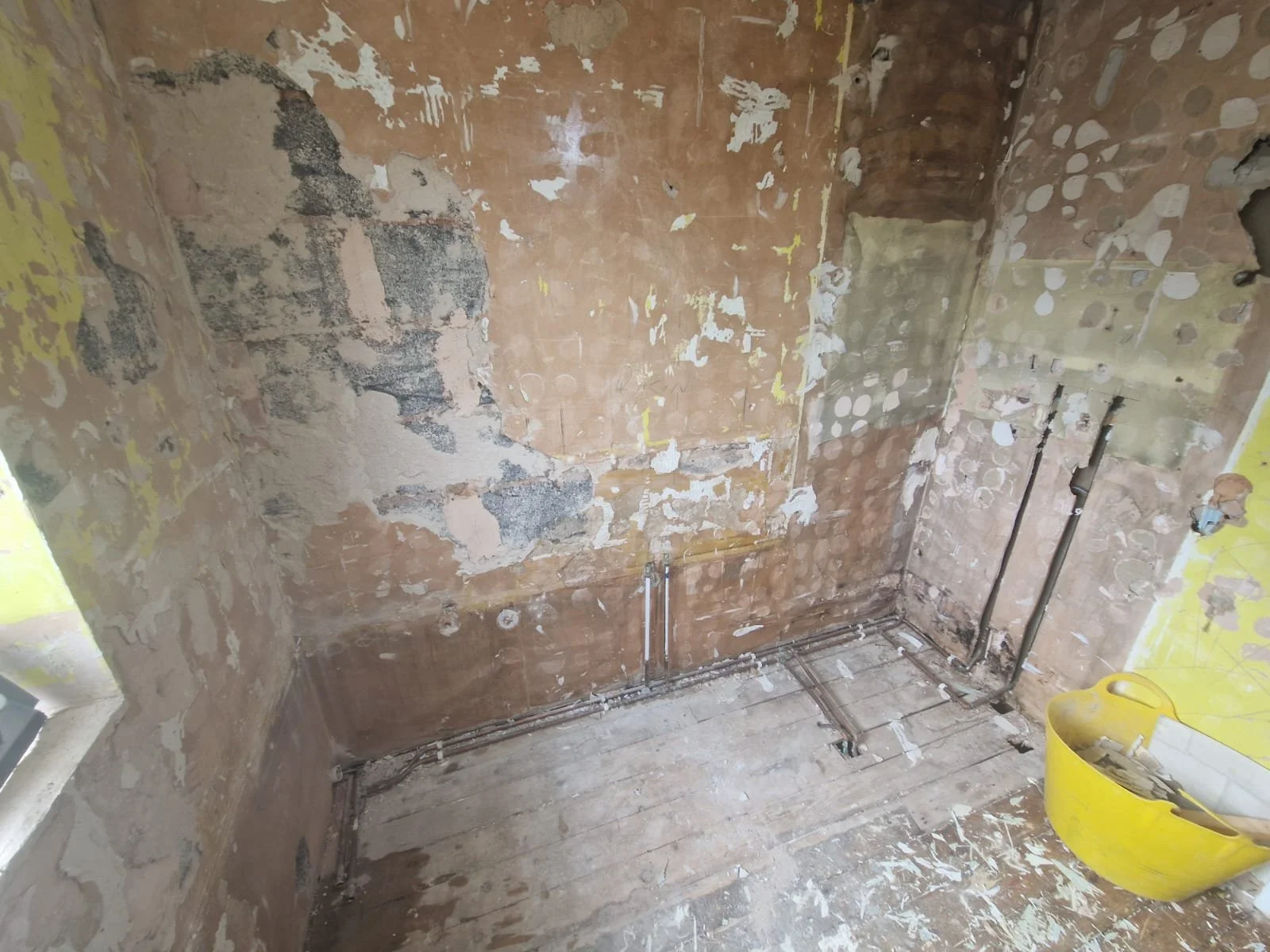 Room under renovation with exposed brick walls, visible pipes, and a yellow bucket on the floor.
