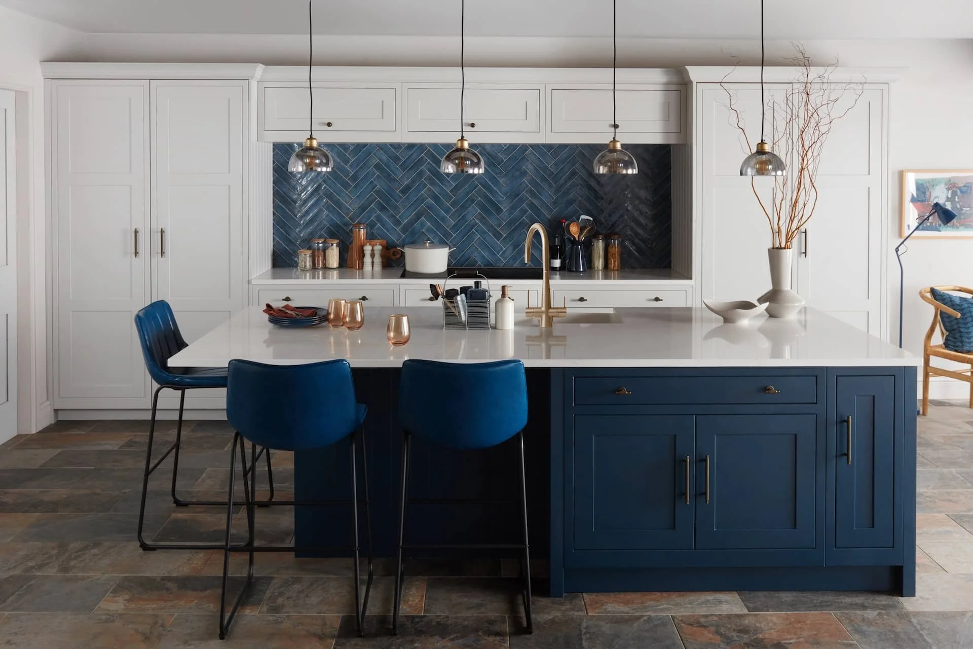 Modern kitchen with navy blue and white color scheme, featuring a large island with navy blue cabinets, white countertop, and three navy blue chairs. The background has white cabinets, a blue herringbone tile backsplash, and pendant lights hanging above the island. Decor includes vases, bowls, and kitchen utensils.