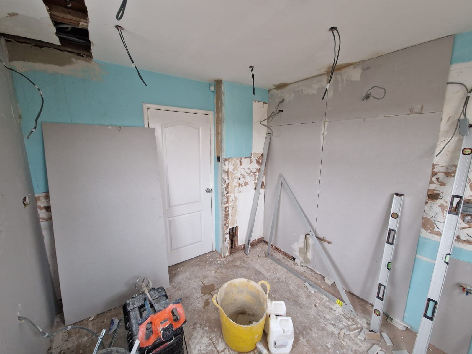 An interior room under renovation with drywall being installed on the walls, electrical wires hanging from the ceiling, construction tools and materials on the floor, and exposed brick and drywall patches.