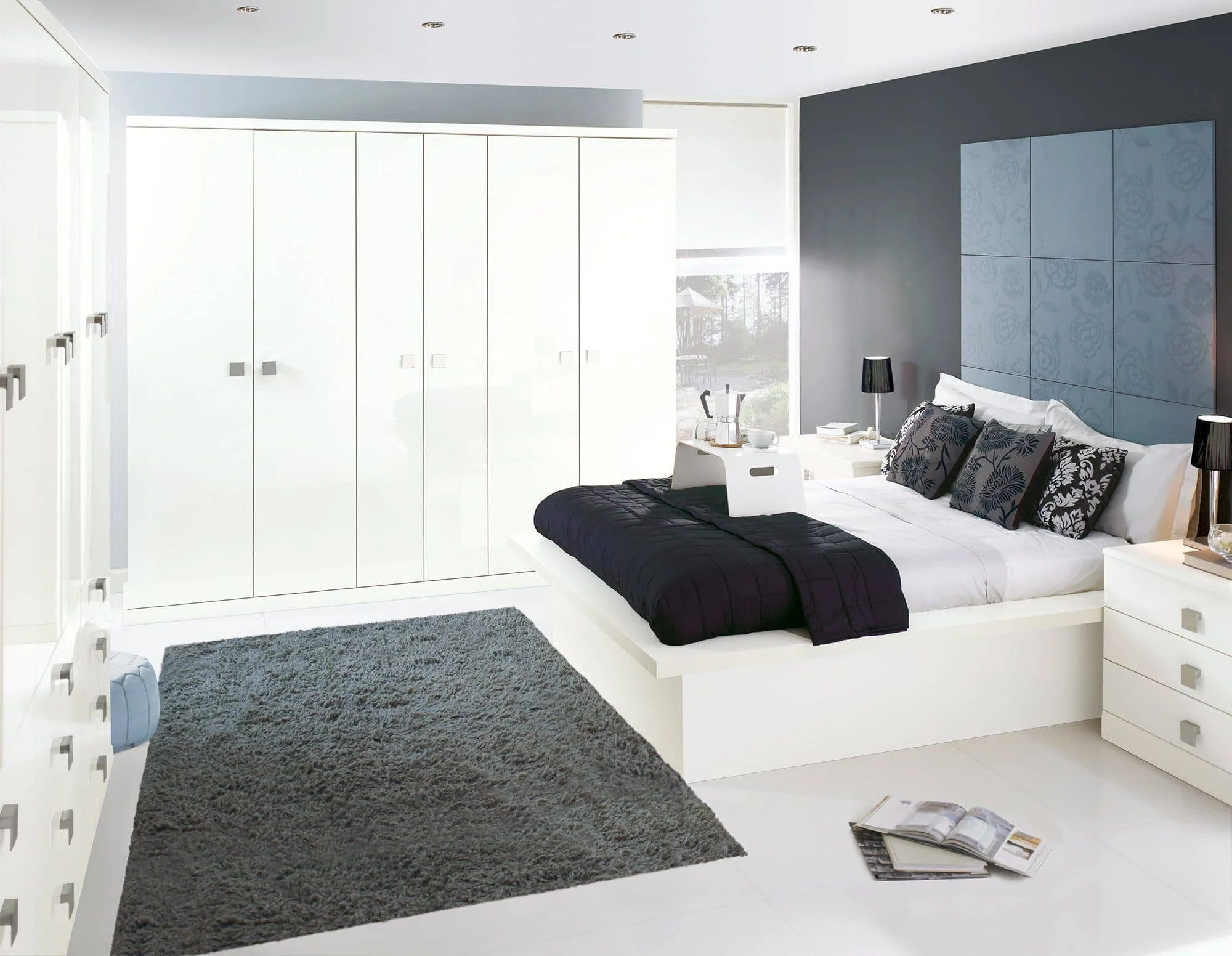 Modern bedroom with white walls, a large white bed with black and patterned pillows, black lamps on white nightstands, a gray rug, a white dresser, and a large white wardrobe. There are magazines on the floor.