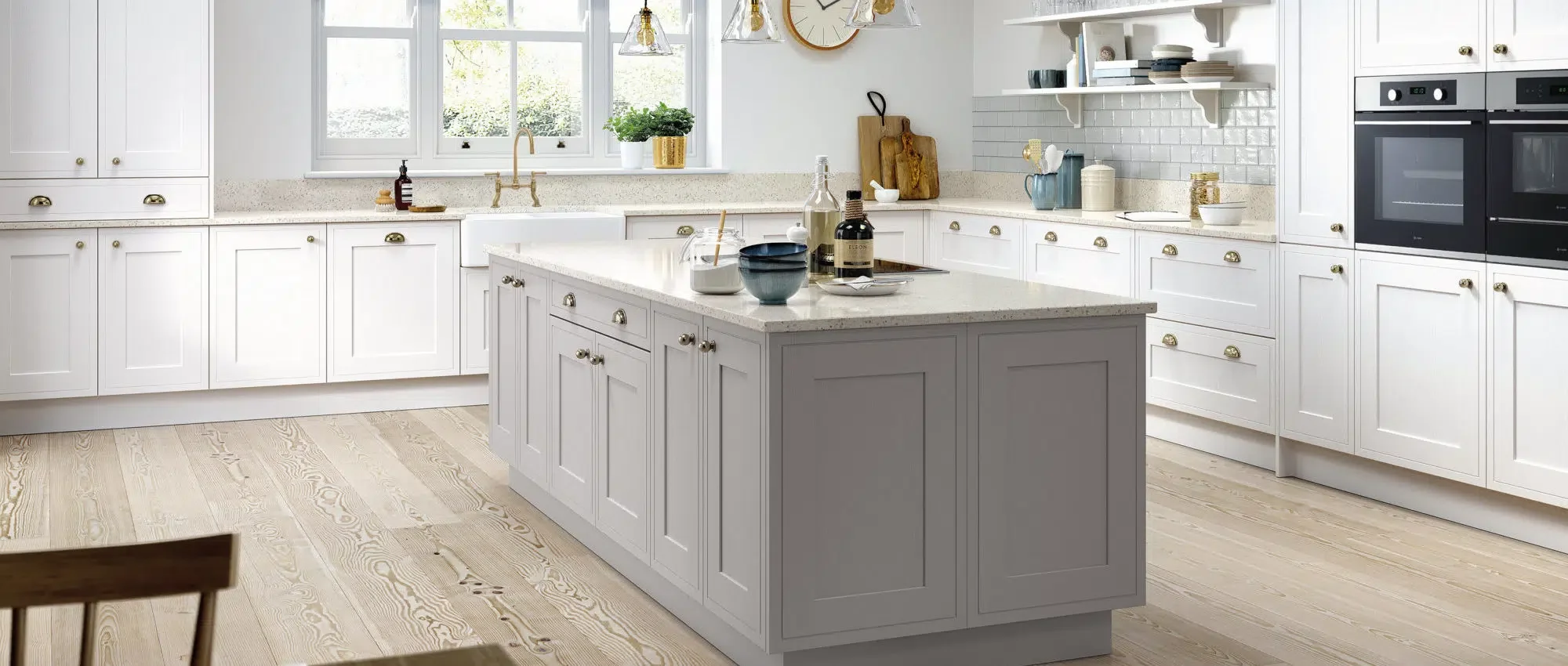 Bright white kitchen with window view, light wood flooring, and central island with dishes and bottles. White cabinetry, open shelves, and built-in oven.