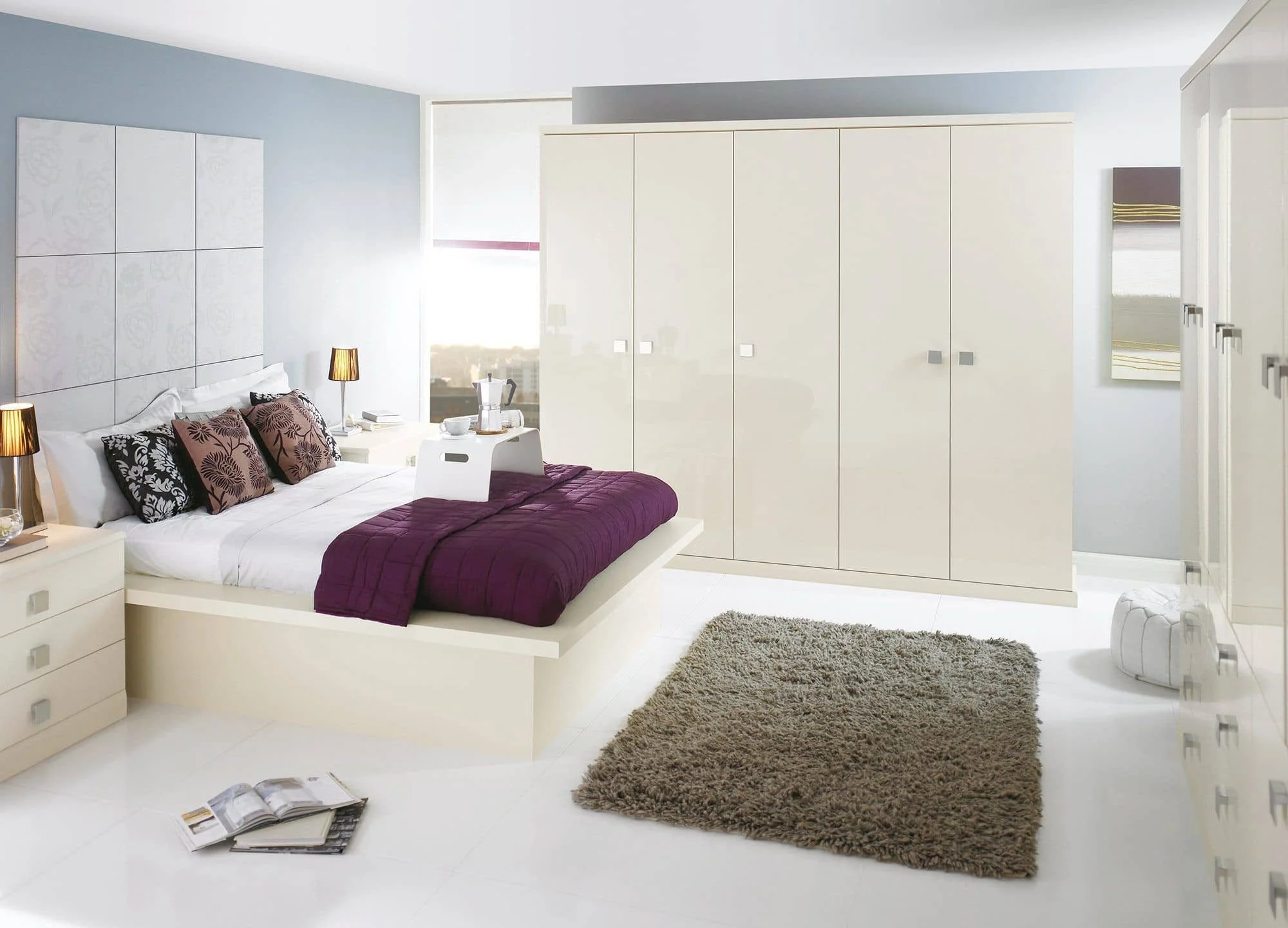 Modern bedroom with white furniture, a bed with purple bedding and patterned pillows, a beige rug, and large wardrobes, sunlight coming through windows.