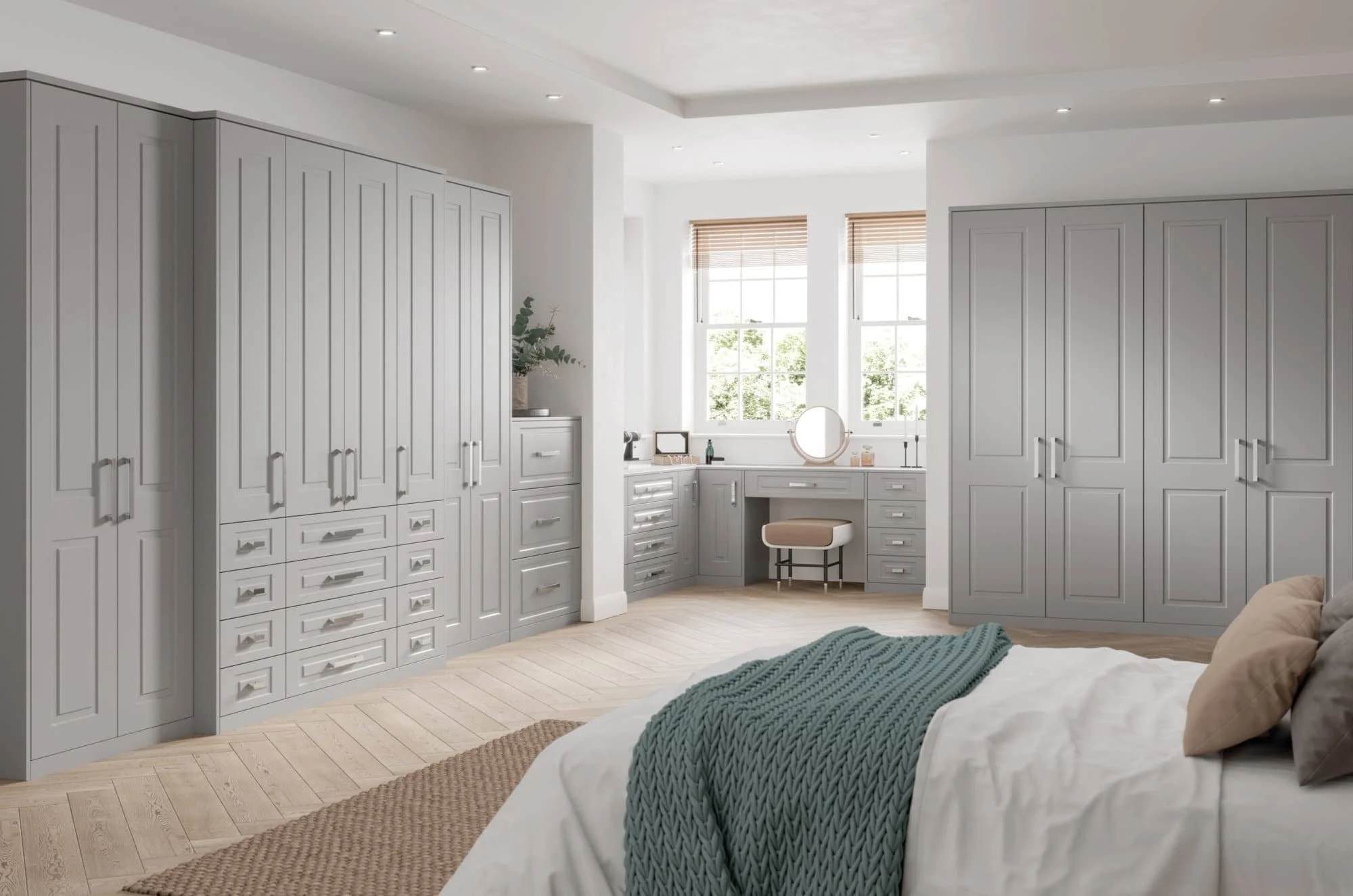 Empty bedroom with a bed, built-in gray wardrobes, and a small vanity desk near windows with blinds, illuminated by recessed ceiling lights.