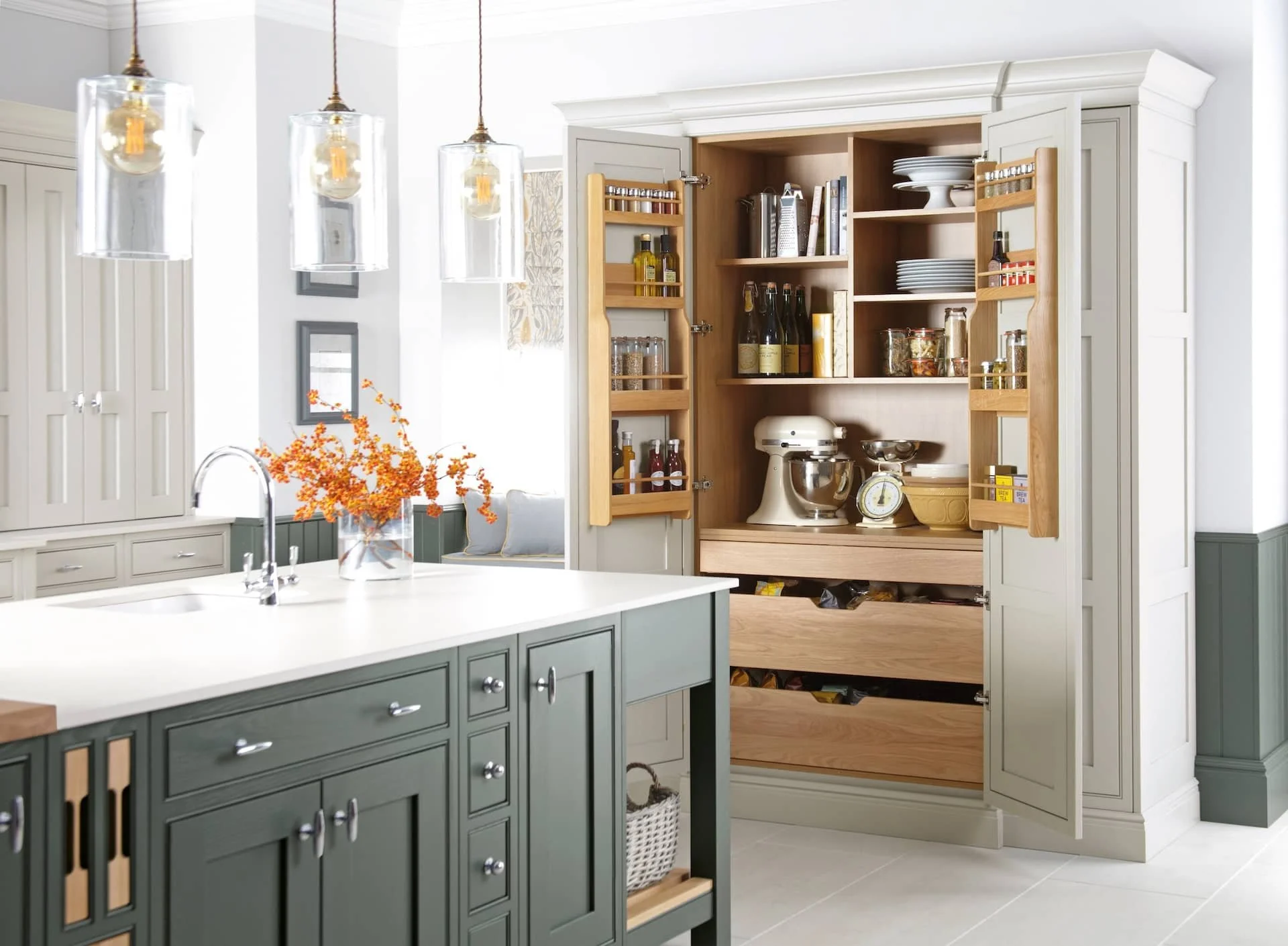 A modern kitchen with white and grey cabinetry, hanging glass pendant lights, and a wooden open pantry containing kitchen appliances, bottles, jars, plates, and bowls. There is a vase with orange flowers on the white kitchen island.