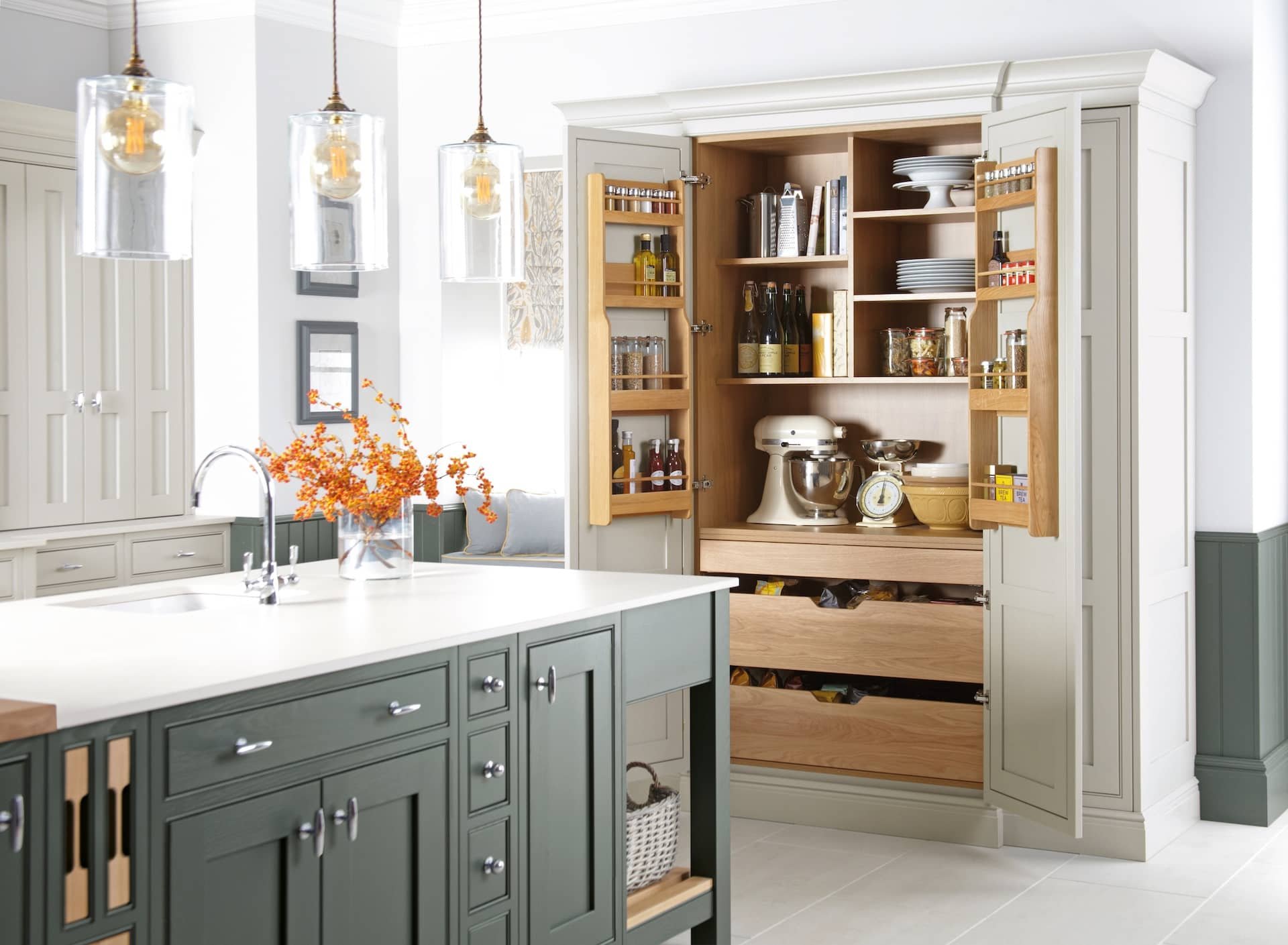 Kitchen with white and gray cabinets, pendant lights, and an open pantry cabinet with shelves, dishes, and bottles. There is a vase with orange berries on the kitchen island.