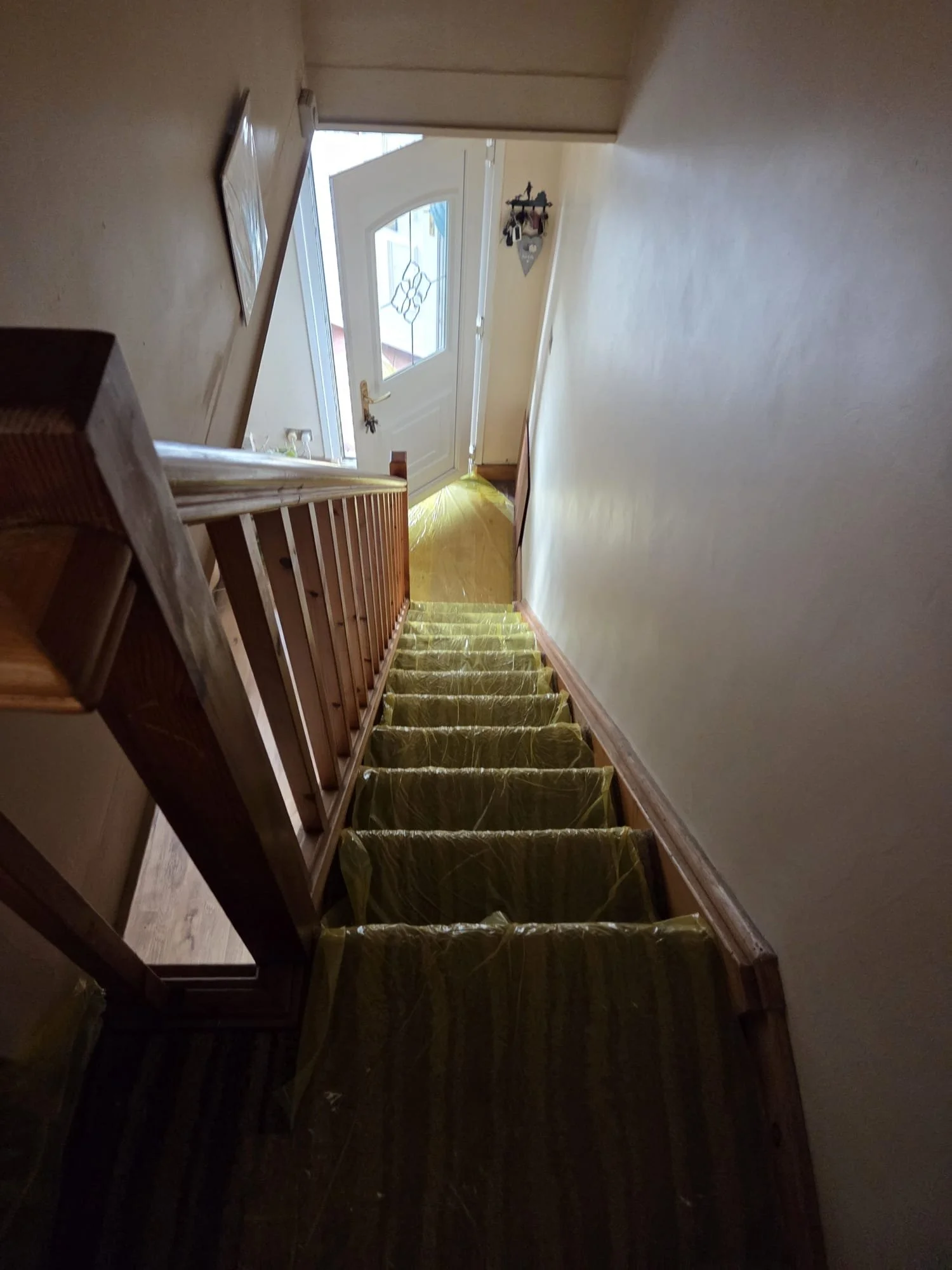 View of a staircase covered with yellow protective plastic, leading down to a front door with a decorative window, in a hallway with white walls and wooden trim.