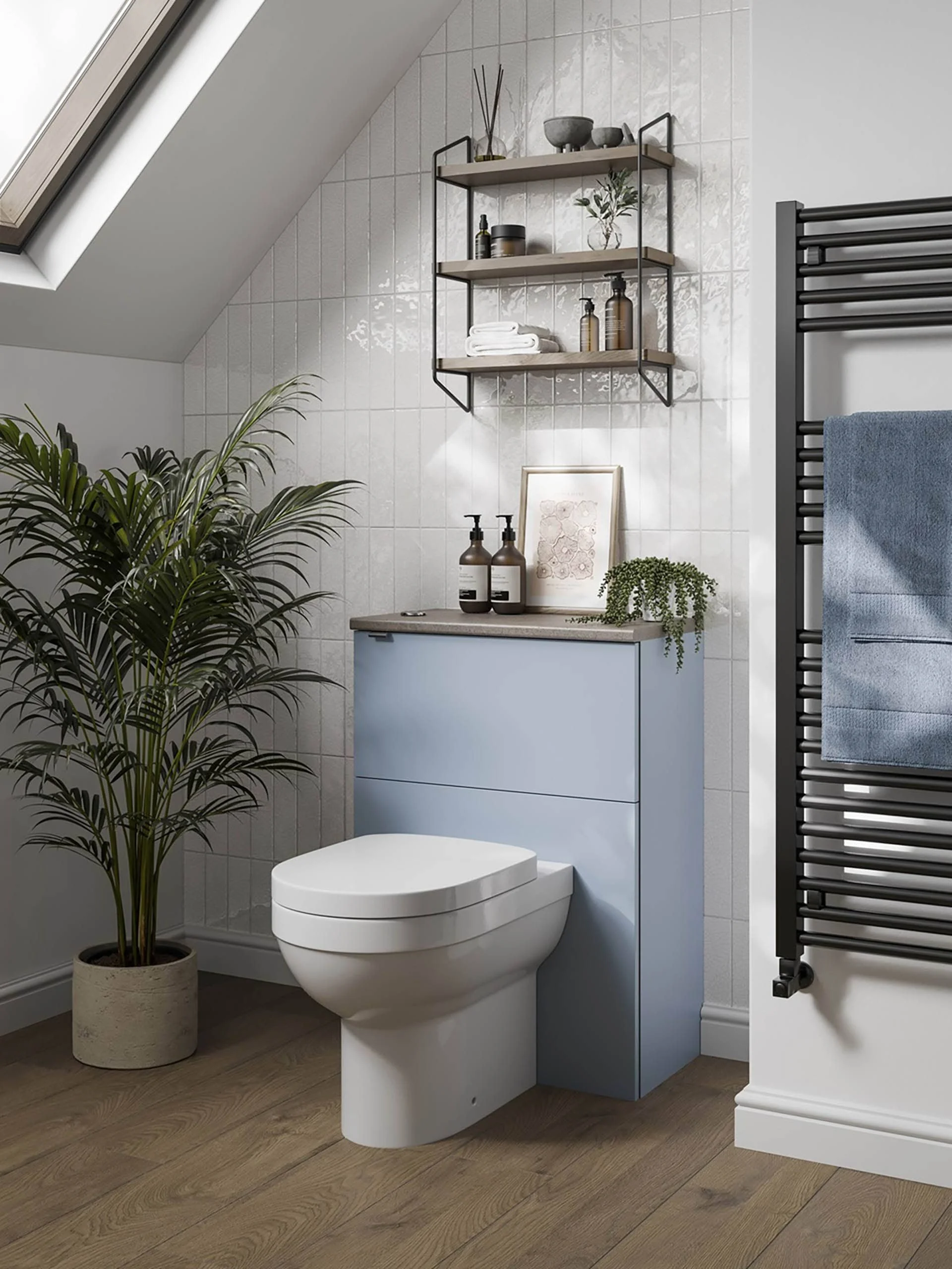 Modern bathroom with a sloped ceiling, skylight window, and white tiled walls. Features a white toilet, a light blue cabinet with a countertop holding two soap dispensers, a framed picture, and green plants. There's a black ladder-style towel rack wi