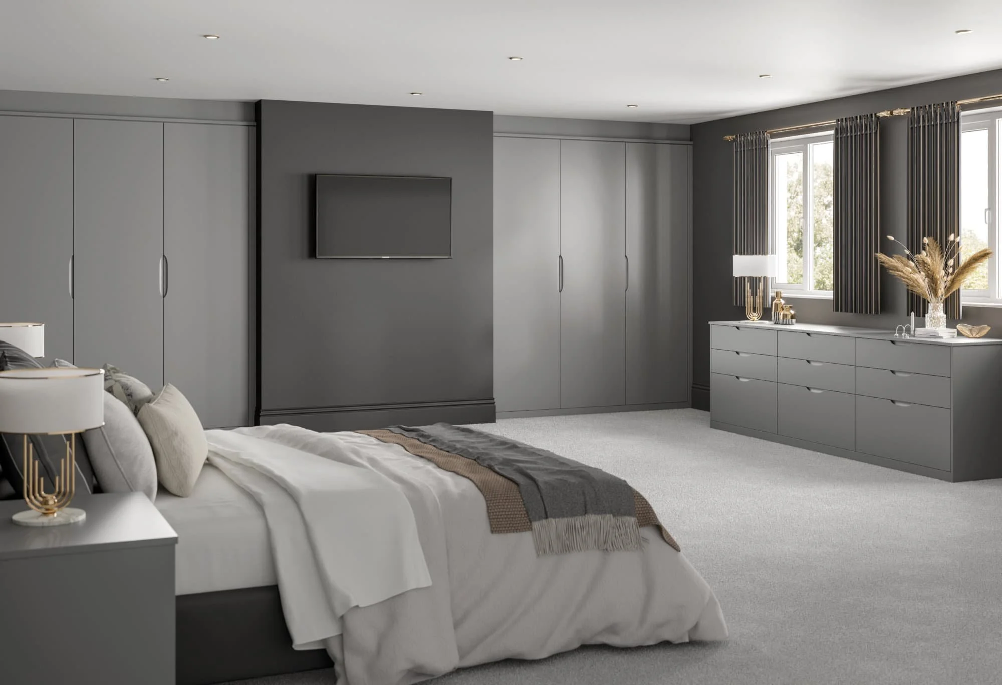 A modern bedroom with gray walls, a large bed with white and beige bedding, gray pillows, and a nightstand with a white lamp. There are built-in gray wardrobes, a wall-mounted TV, and a dresser with decorative items, including a vase with dried plant