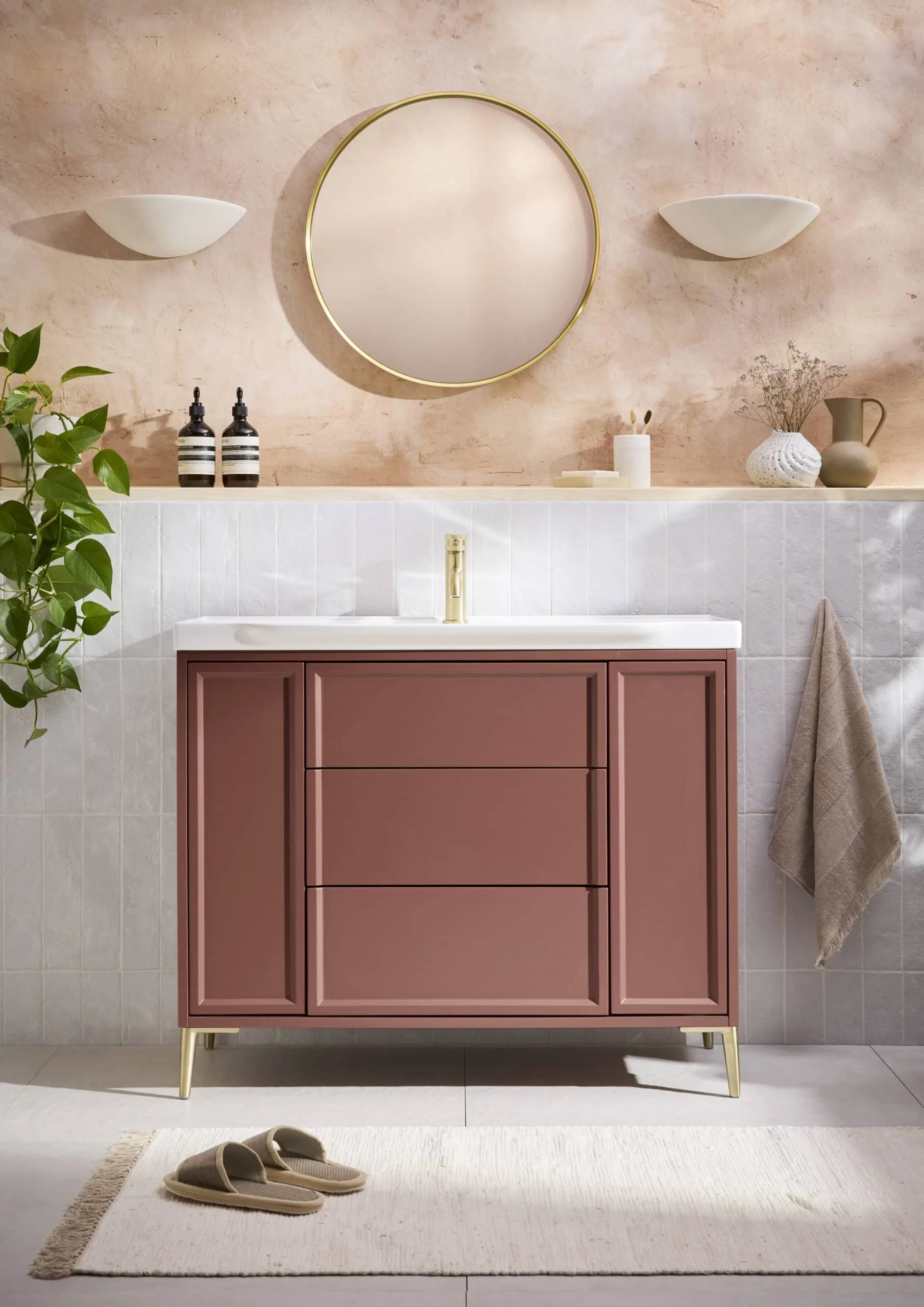 A modern bathroom with a pink vanity, a white countertop, and gold accents. There is a large round mirror above the vanity, and decorative items on the shelf including vases and bottles. A pair of slippers is on a small white rug on the floor next to
