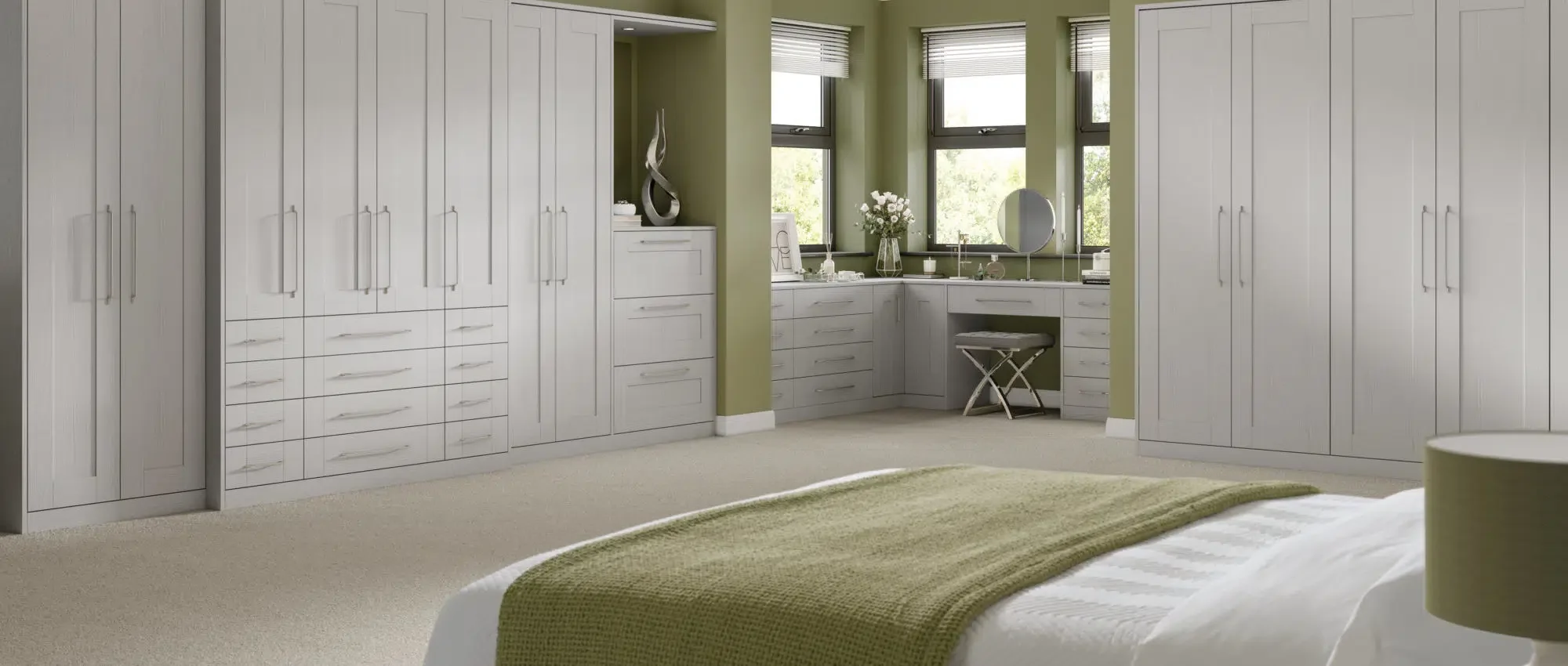 A bedroom with built-in white wardrobes lining the walls, a bed with a green blanket, and a corner vanity area with a window, a mirror, and a vase of white flowers.