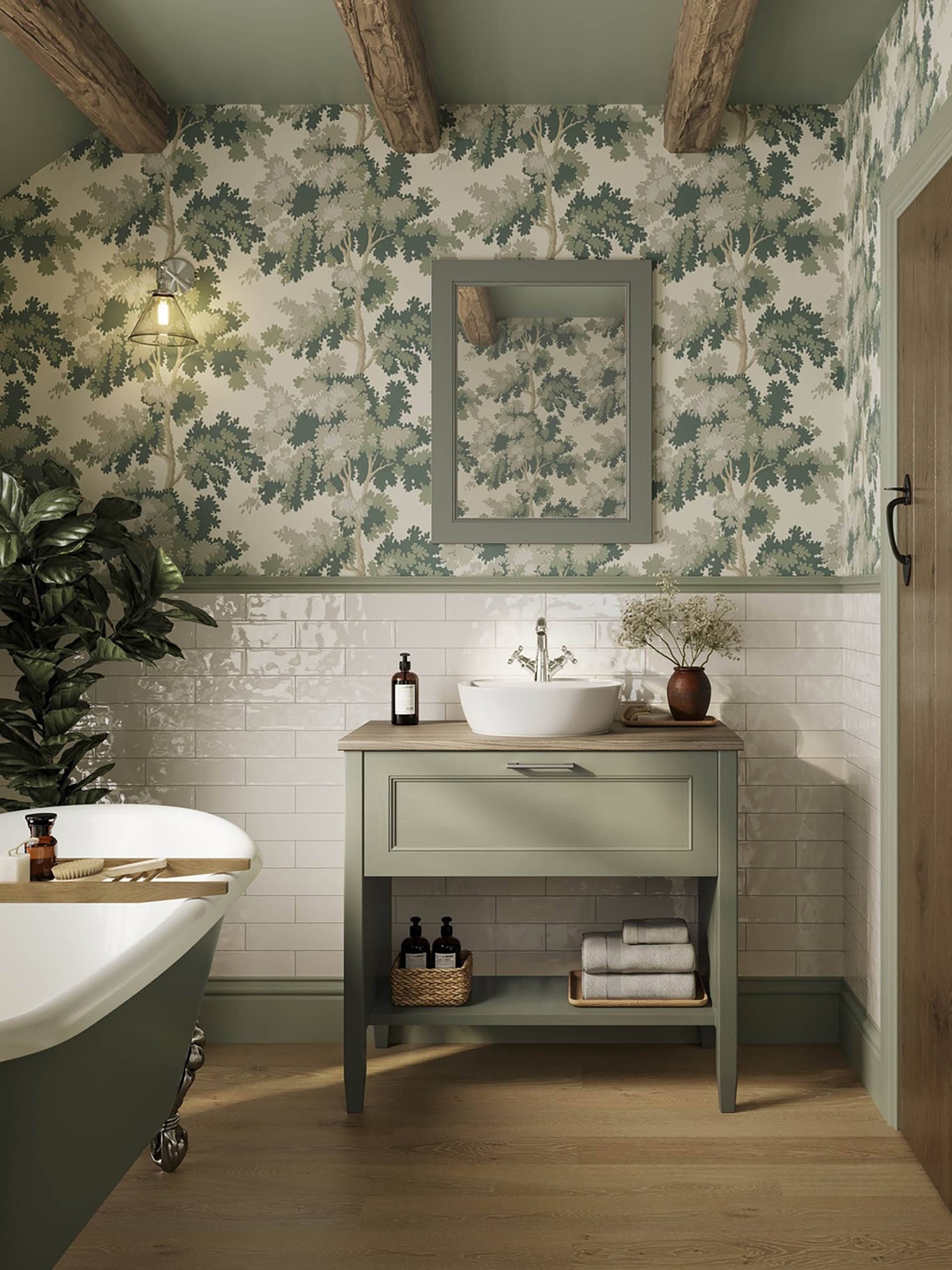 A bathroom with green and white botanical wallpaper, a small green vanity with a white vessel sink, a mirror above, and a bathtub to the left. Decor includes a potted plant, rolled towels, and toiletry bottles.