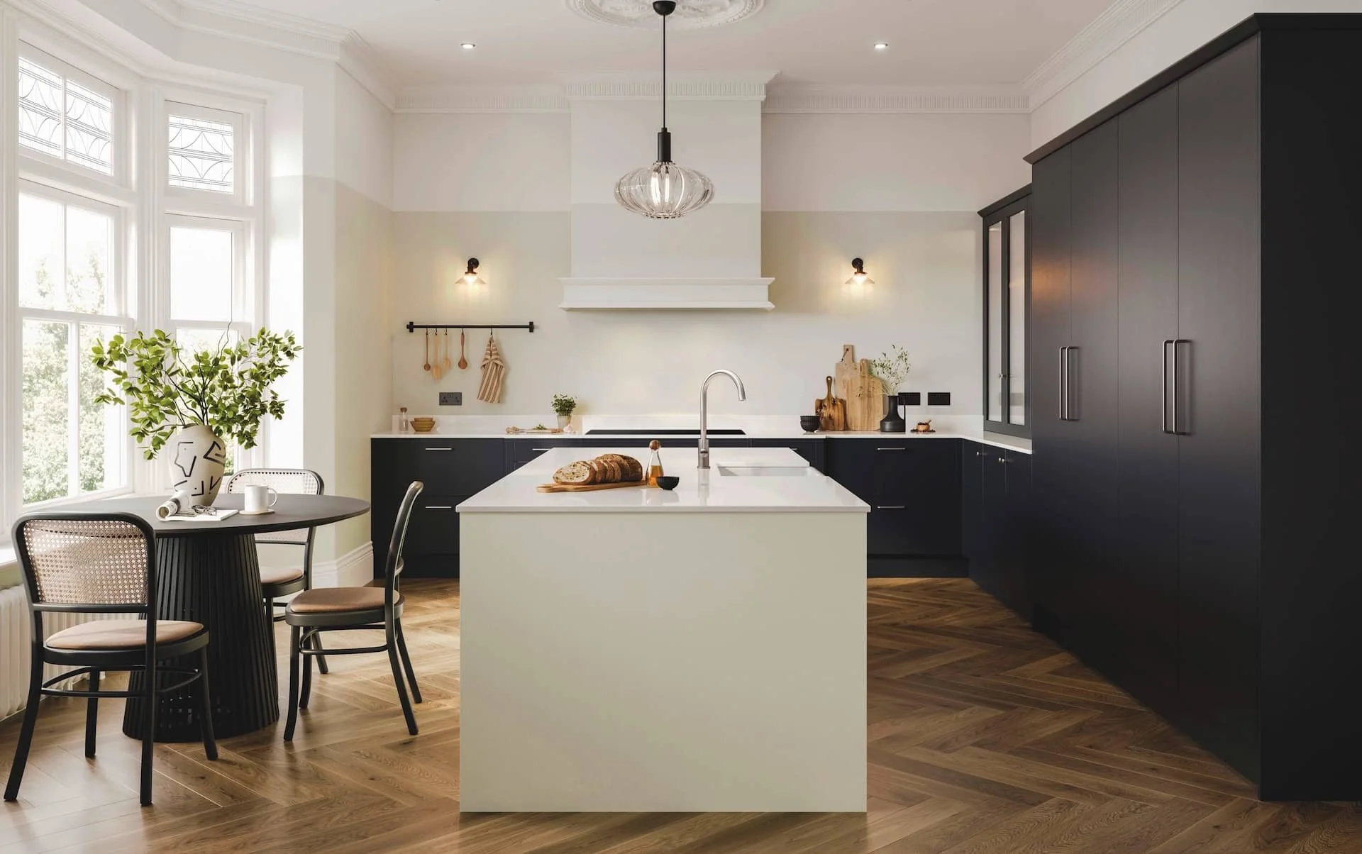 Modern kitchen with white island, black cabinets, wooden flooring, and large windows. Dining table with flowers and chairs to the left.