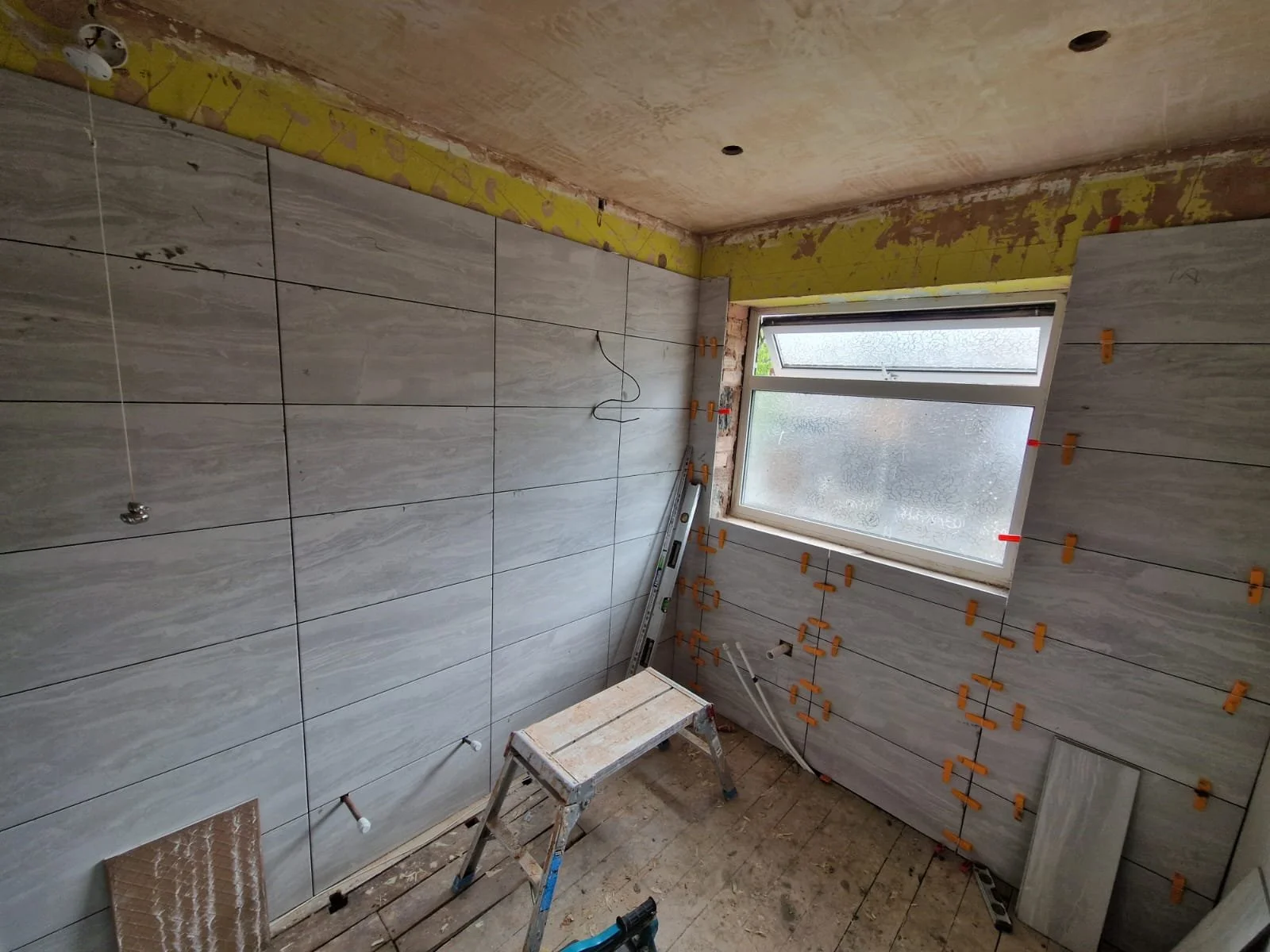 A room under construction with large gray tiles being installed on the walls, a window with frosted glass, and construction tools and materials on the floor.
