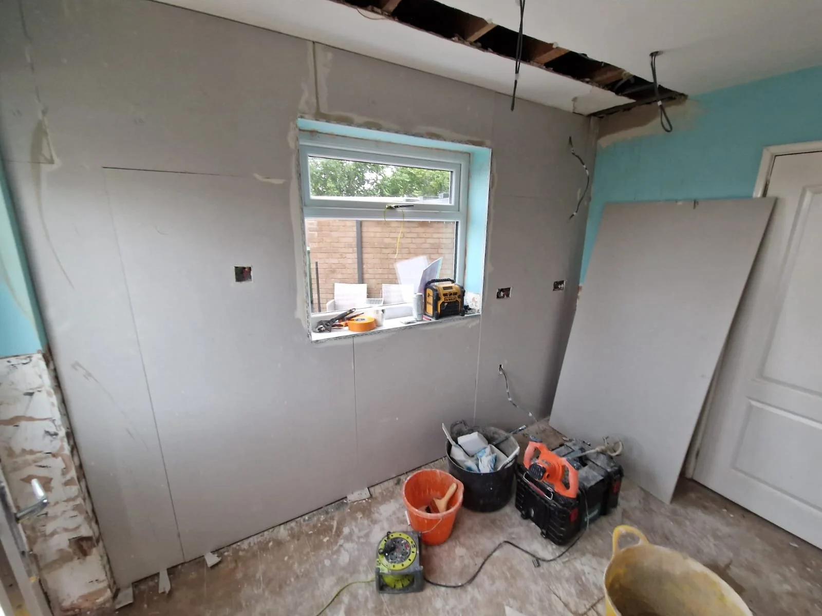 Room under renovation with partially installed drywall, tools, and construction materials on the floor, window revealing outdoor brick wall and chairs, exposed ceiling with hanging wires.