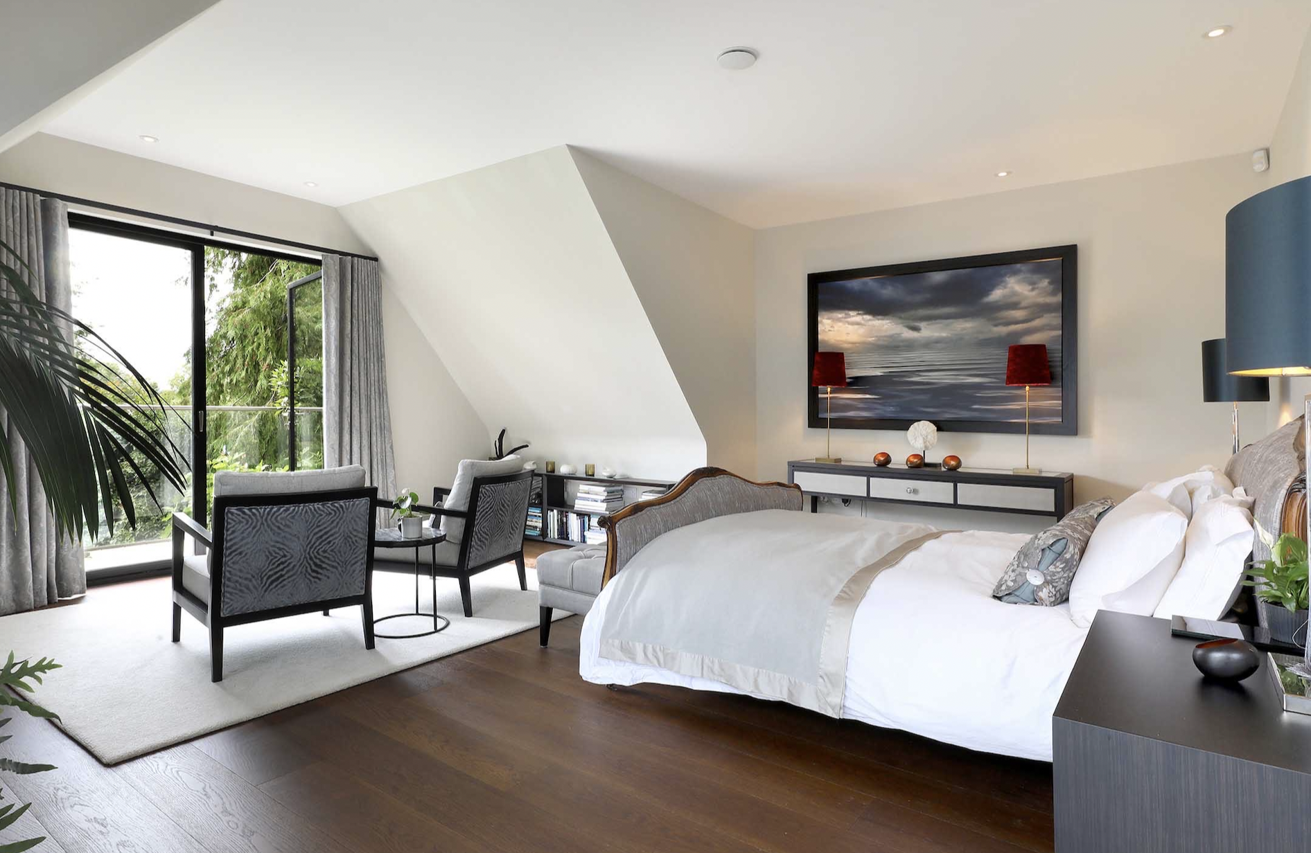 Modern bedroom with large sliding glass door opening to greenery, a sitting area with two chairs and small table, a white bed with decorative pillows, a dark cabinet with a landscape photo above, and various lamps and plants.