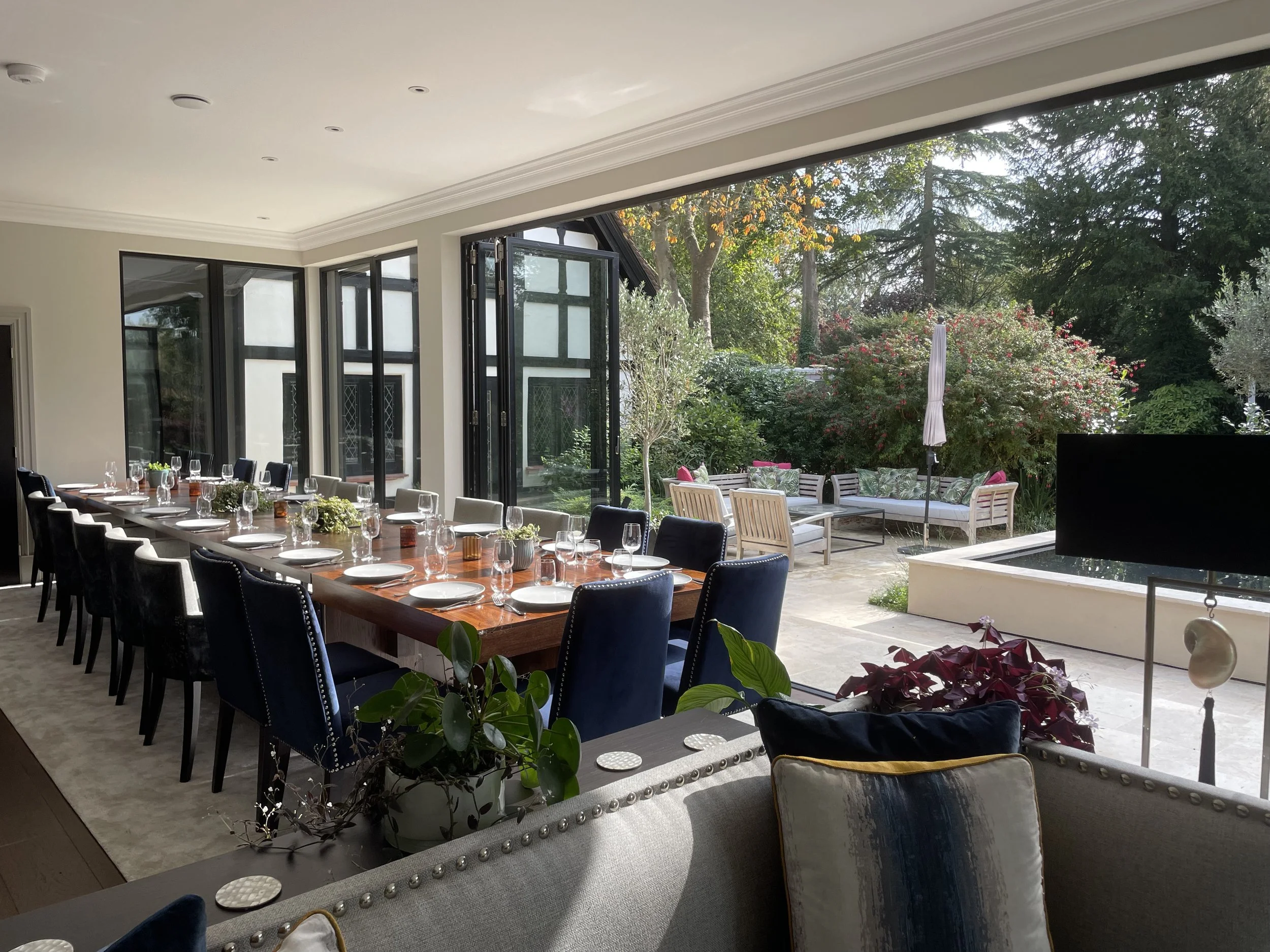 A dining room with a long wooden table set with plates, glasses, and small centerpieces. Large glass doors open to a patio with outdoor seating surrounded by greenery and trees.