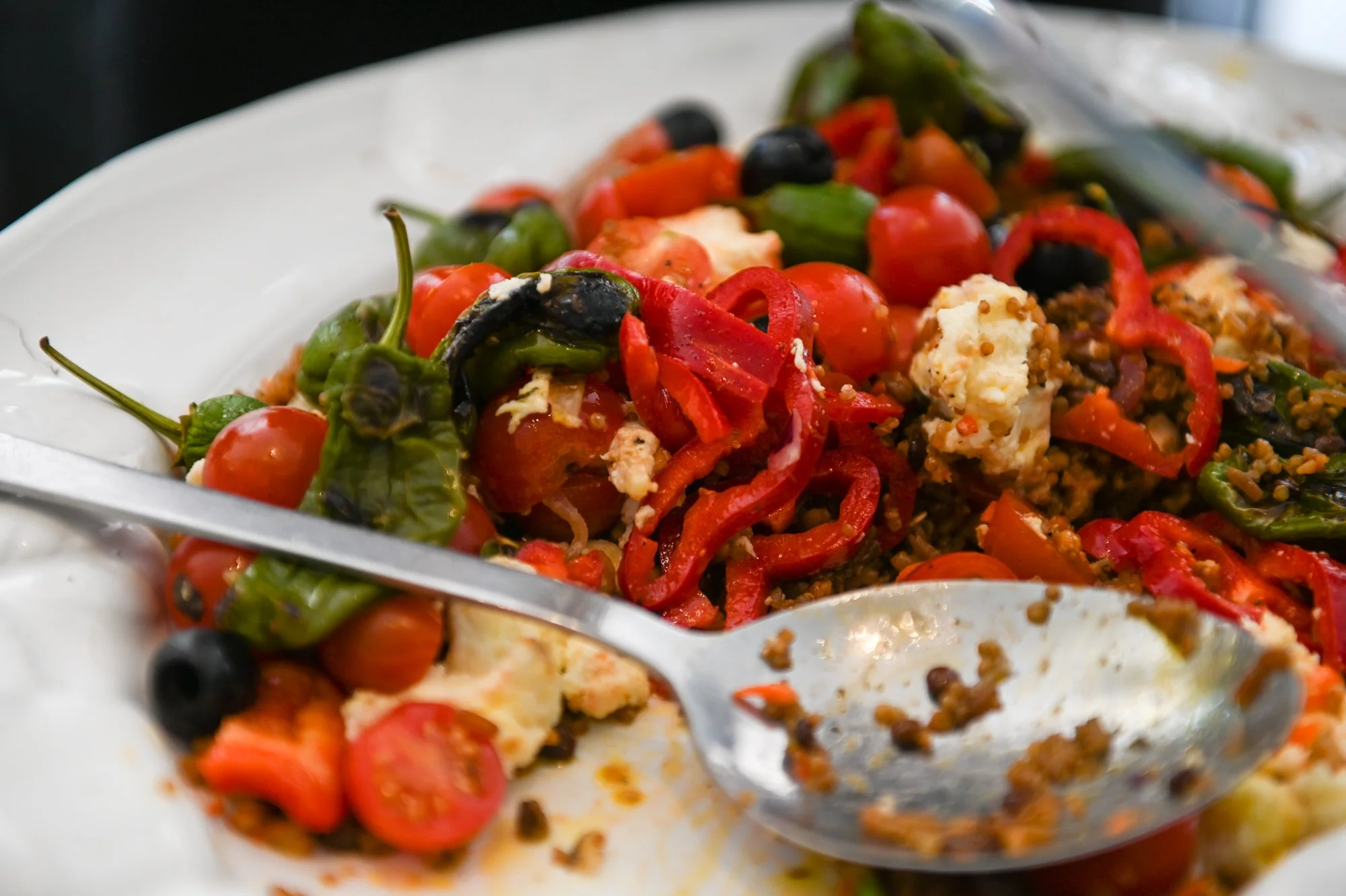 Close-up of a colorful salad with cherry tomatoes, red and green bell peppers, black olives, feta cheese, and cooked ground meat, with a silver serving spoon resting in the bowl.