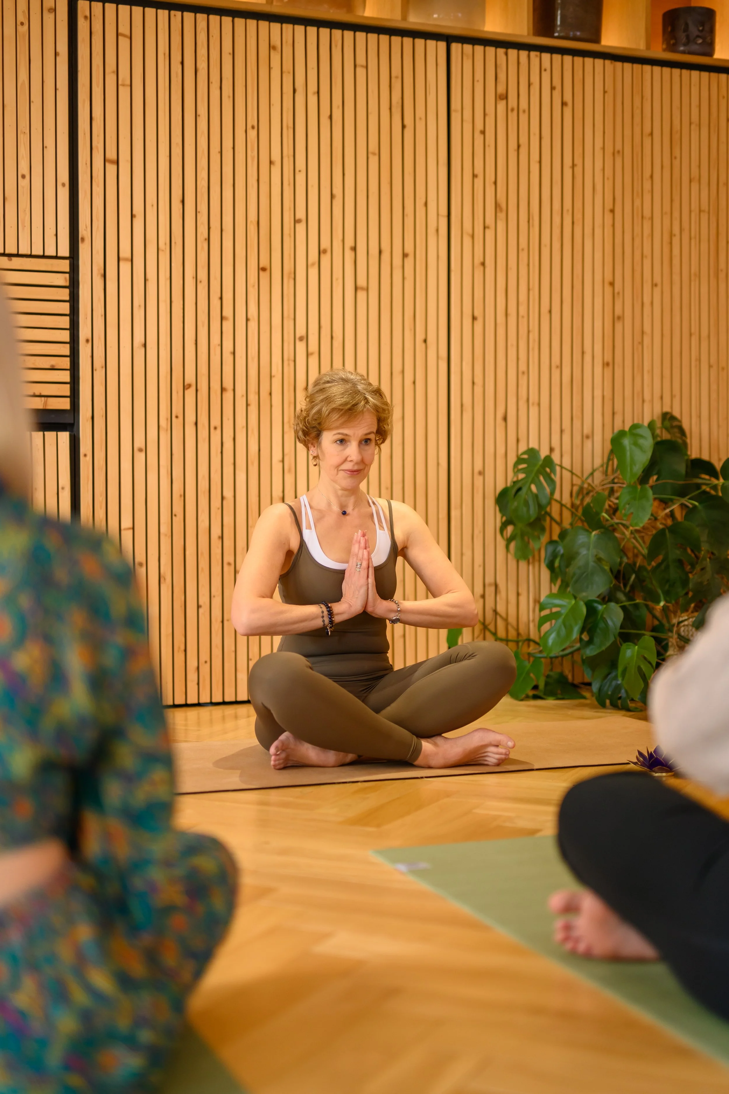A woman practicing yoga or meditation in a studio with wooden walls, sitting cross-legged on a yoga mat with her hands in prayer position, leading a class.
