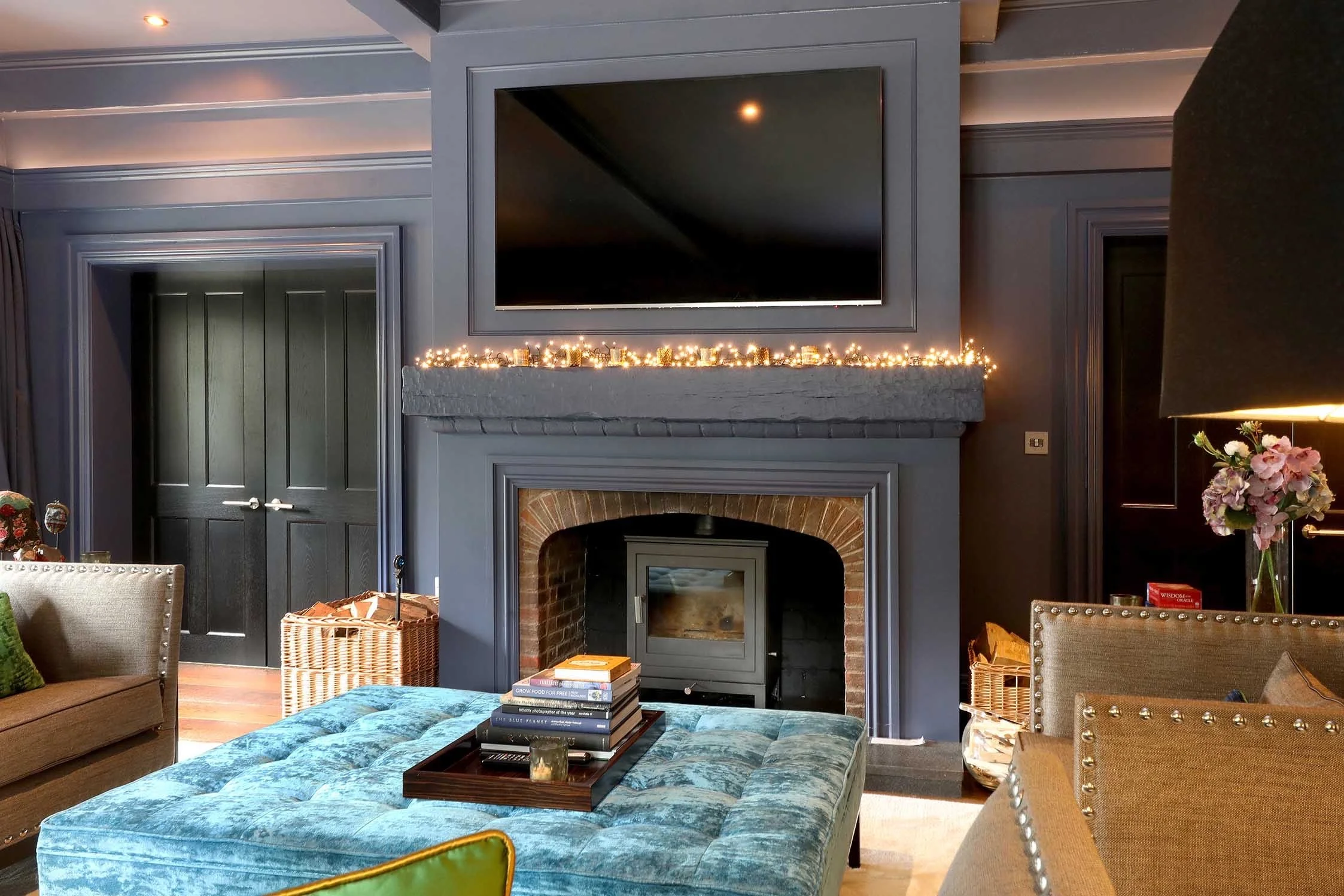 Living room with blue painted walls, a fireplace with a wood stove, stacks of books on a brown tray, a large flat-screen TV above the fireplace, and a tufted ottoman in the center. There are various chairs and a vase of pink flowers on a side table.