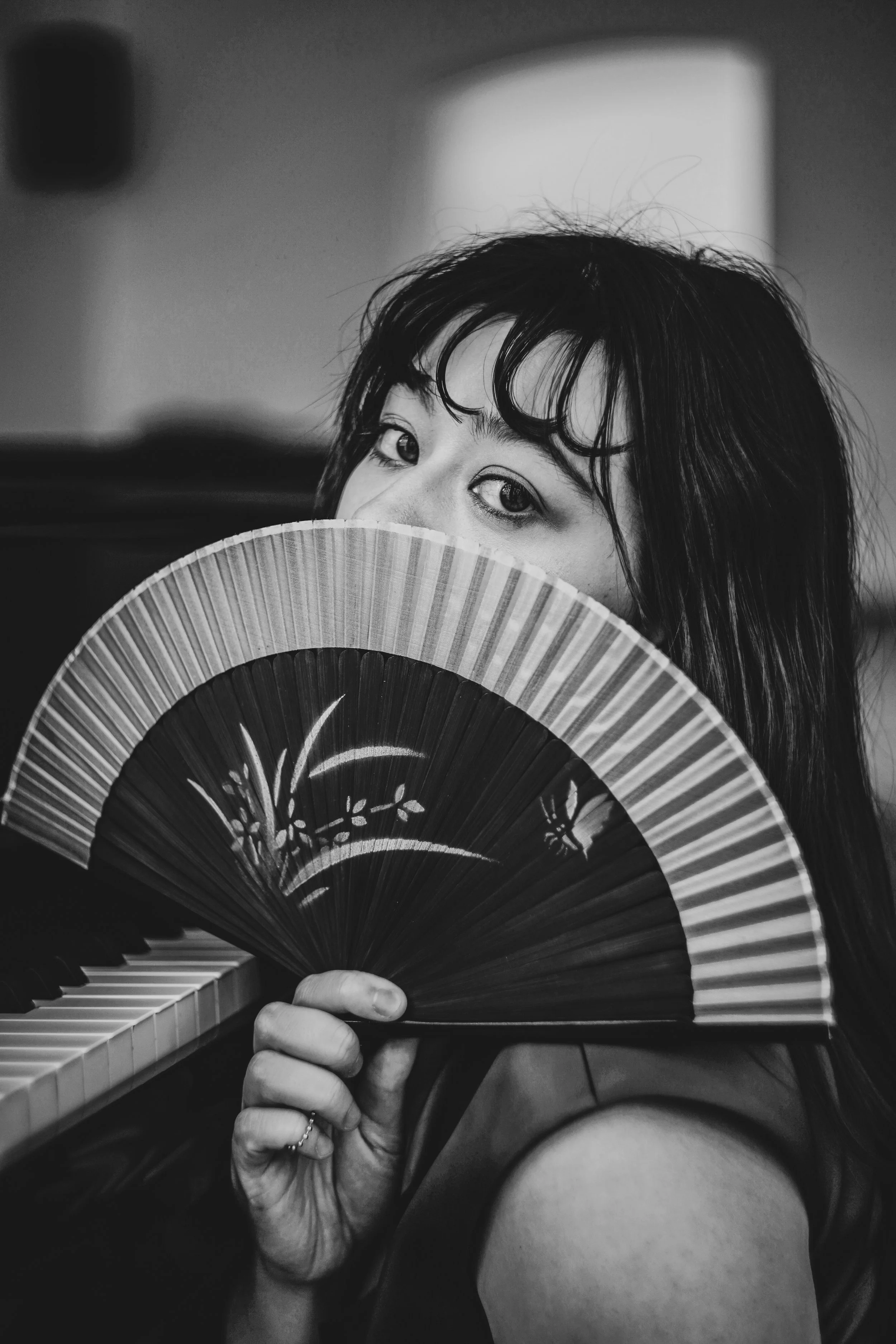 A woman with wet hair holding a decorated hand fan partially covering her face, looking at the camera, with a piano keyboard visible in the foreground.