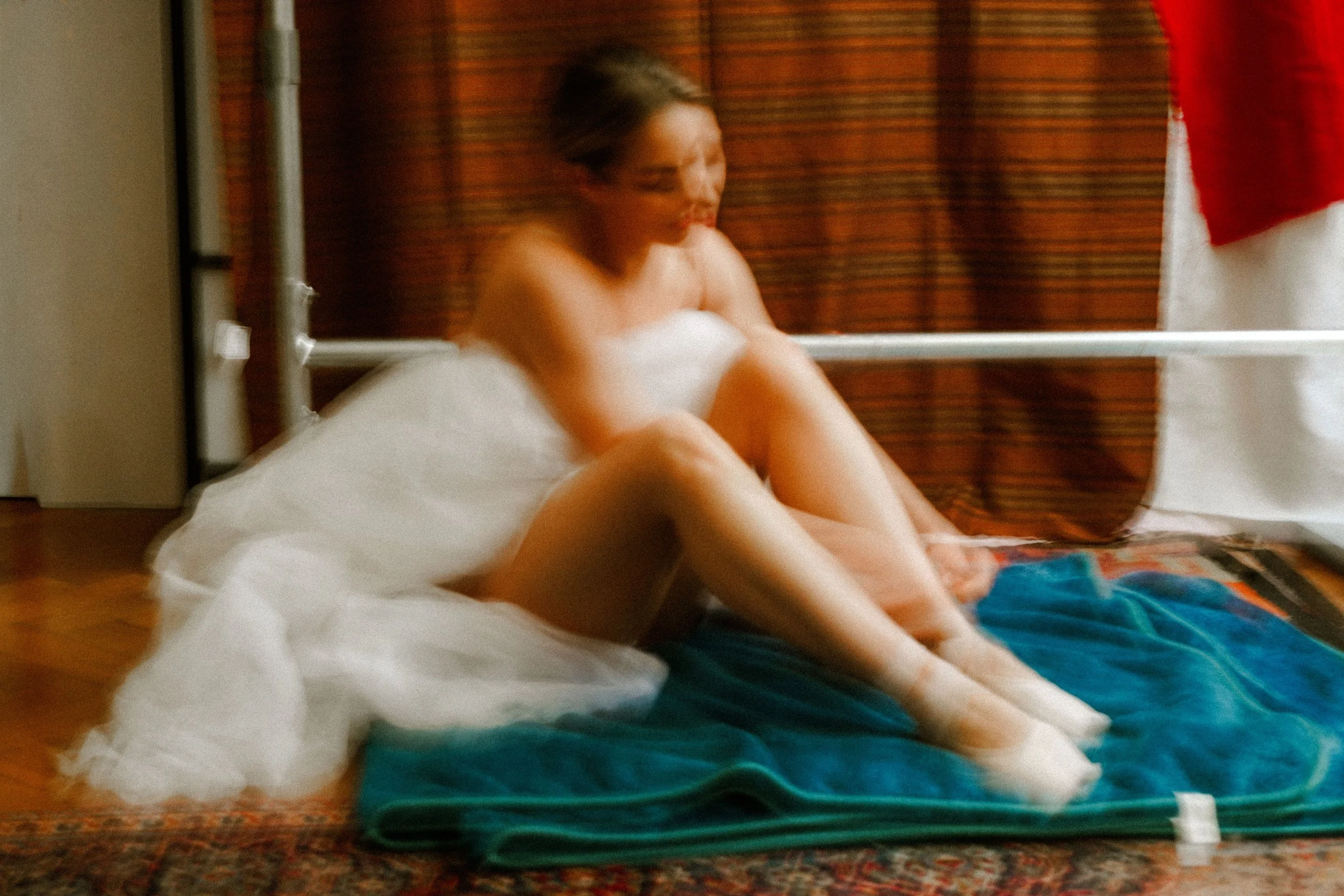 A woman in a white dress sitting on a carpeted floor with a colorful rug, with a blurred background of curtains and a white wall.