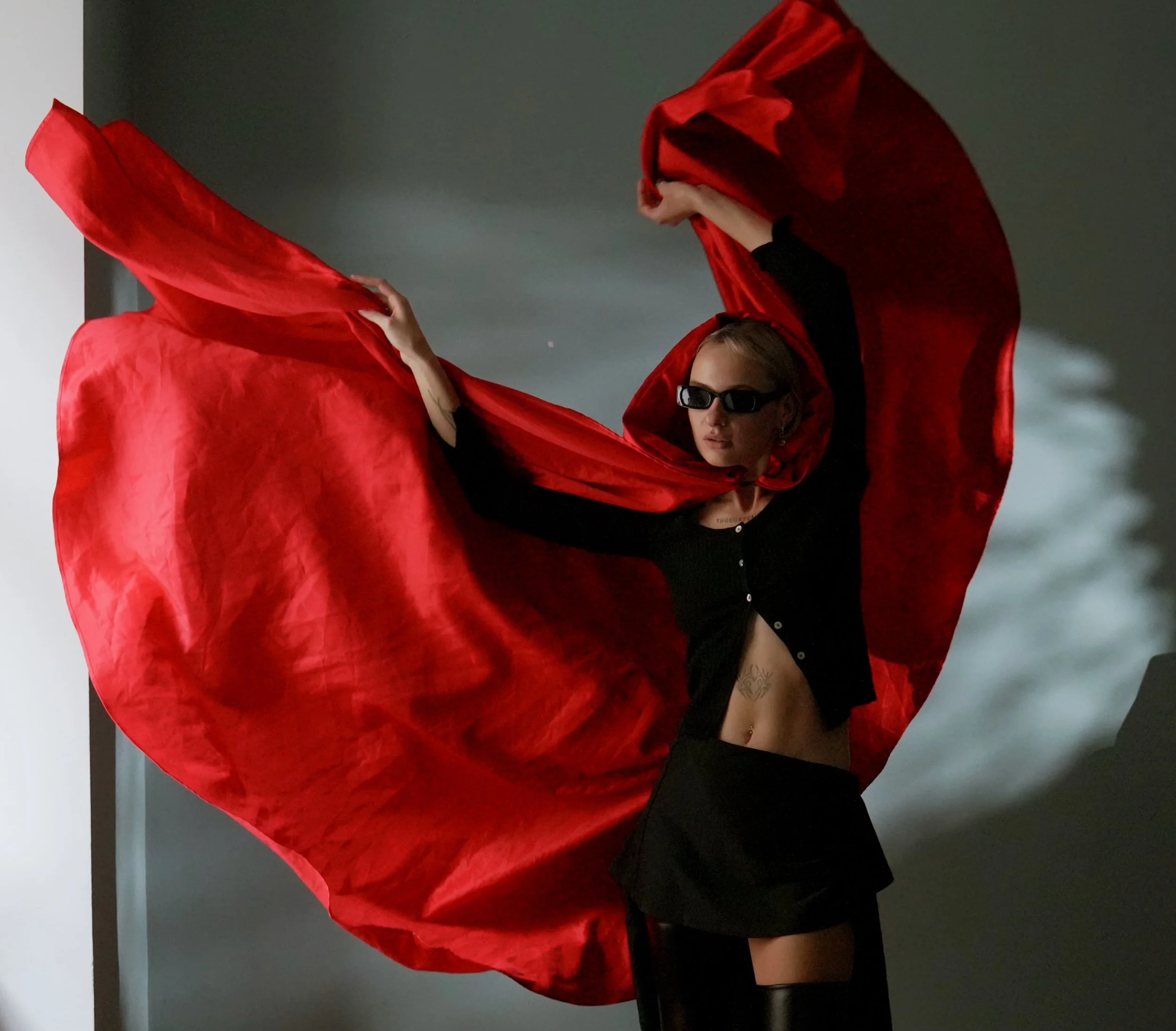 A woman wearing sunglasses and a black outfit holding and waving a large red fabric in front of a plain background.