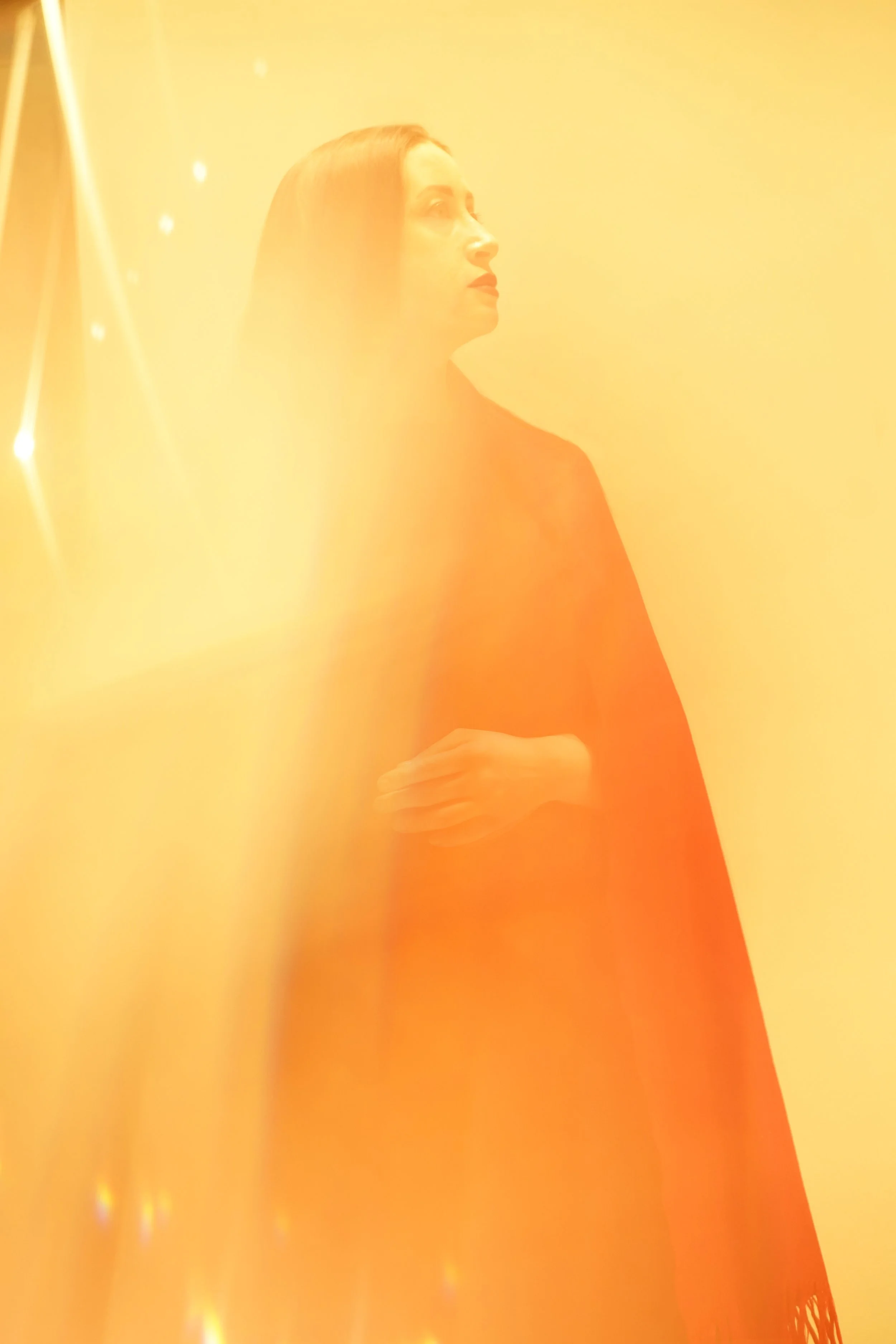 A woman with long hair, dressed in dark clothing, standing against a yellow background, illuminated with warm, golden light.