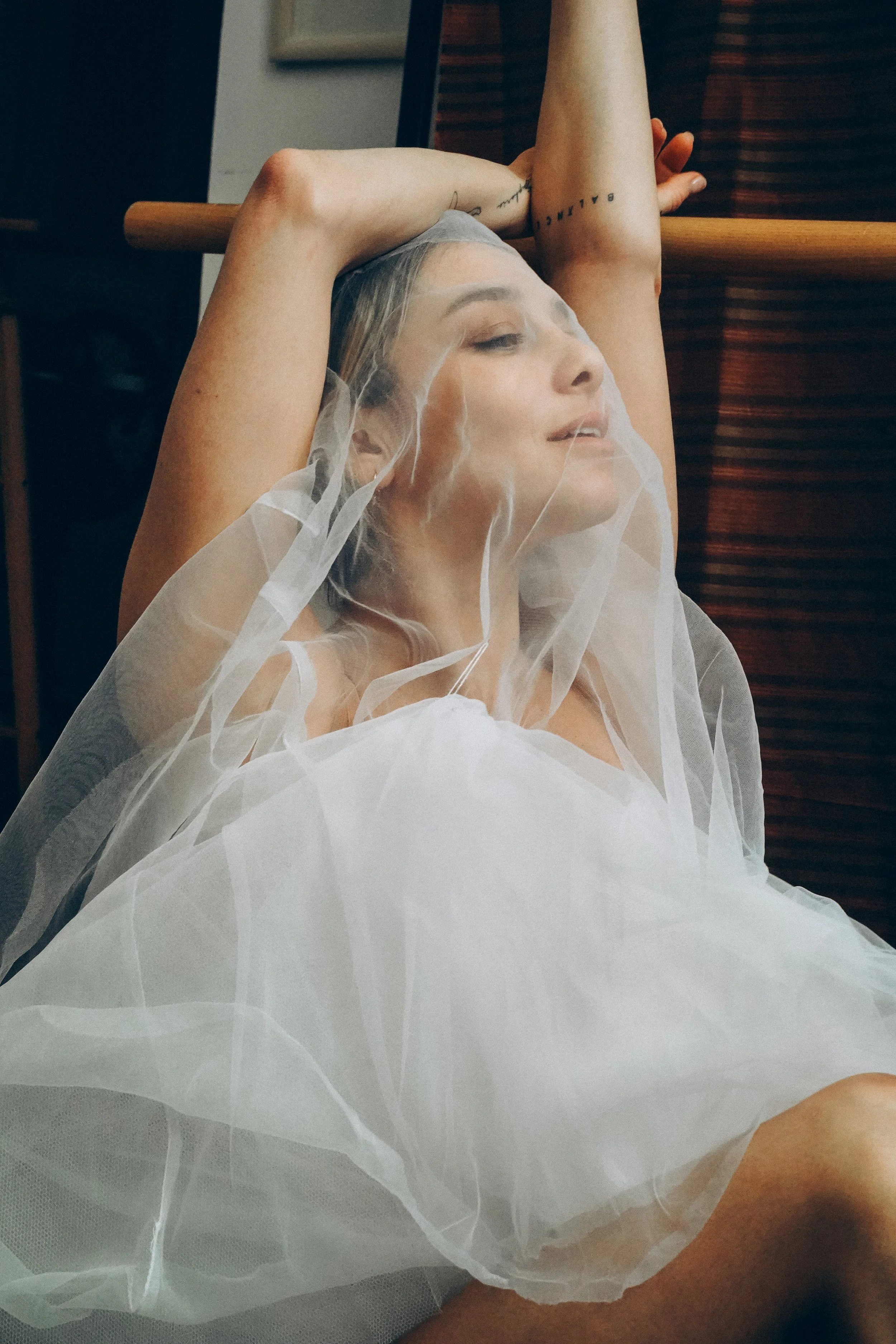 A woman with blonde hair partially covered by a sheer veil, resting with her eyes closed, arm raised above her head, in a relaxed pose.
