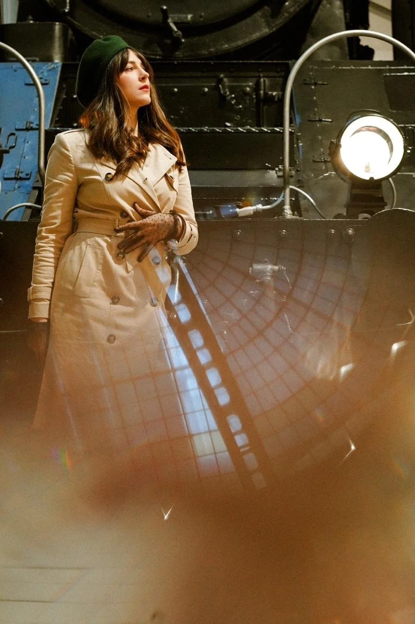 A woman in a beige trench coat, green beret, and brown gloves standing in front of vintage black and blue train machinery with a large spotlight.