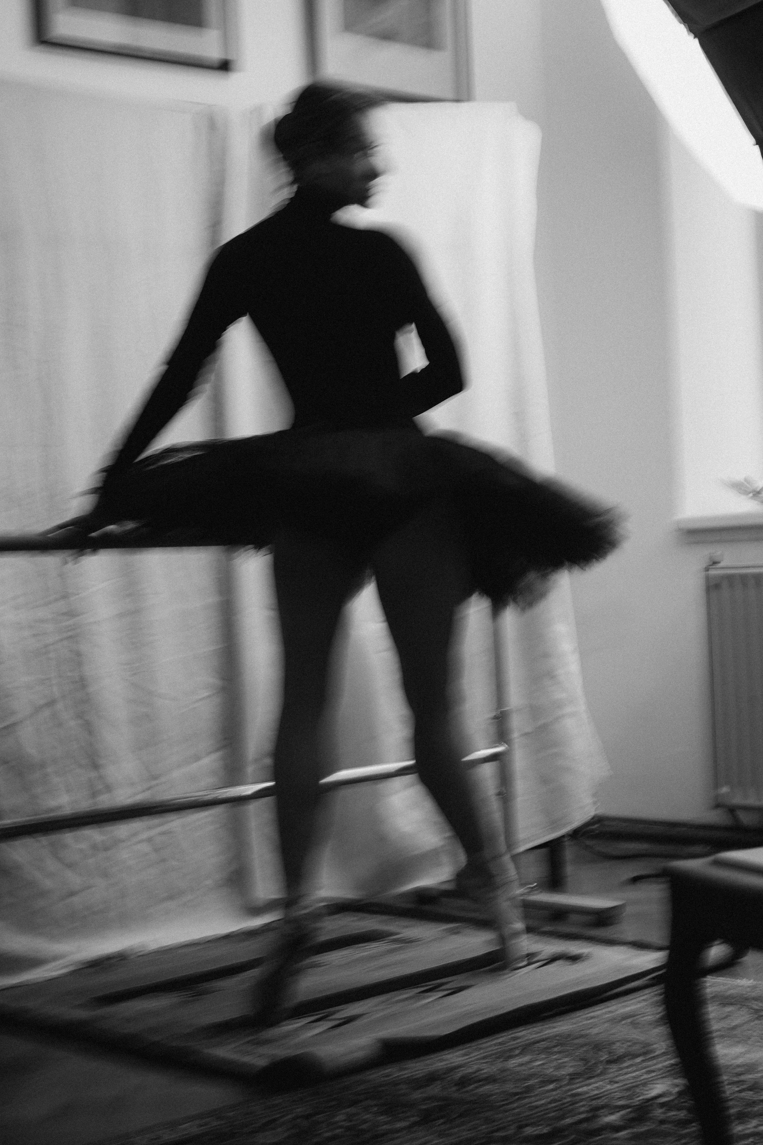 Black-and-white photo of a ballet dancer in a tutu standing on a platform in a studio, with a blurred background and a lighting reflector.