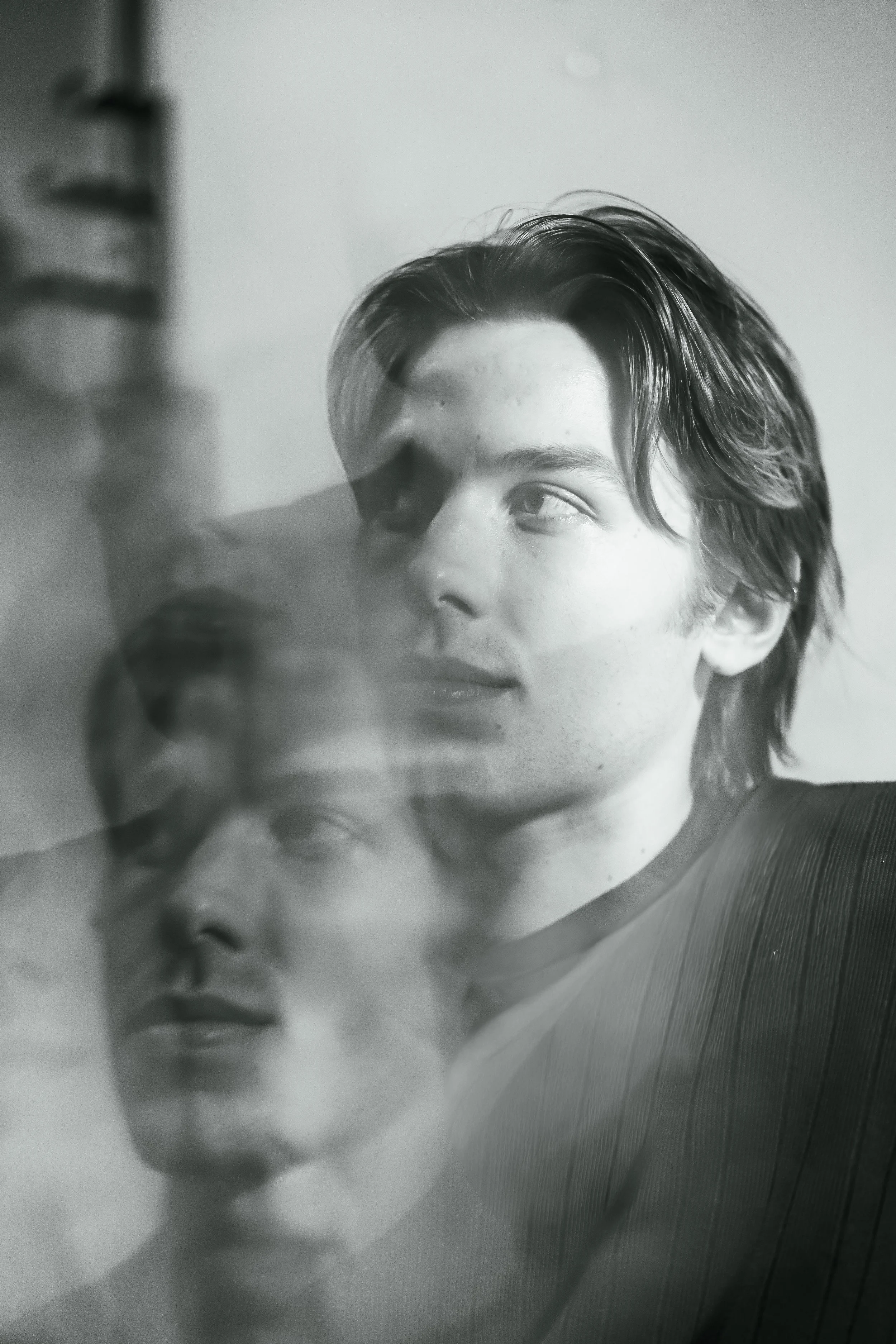 Black and white photograph of a young man with medium-length hair, with a superimposed, ghostly reflection of his face on the left side of the image.