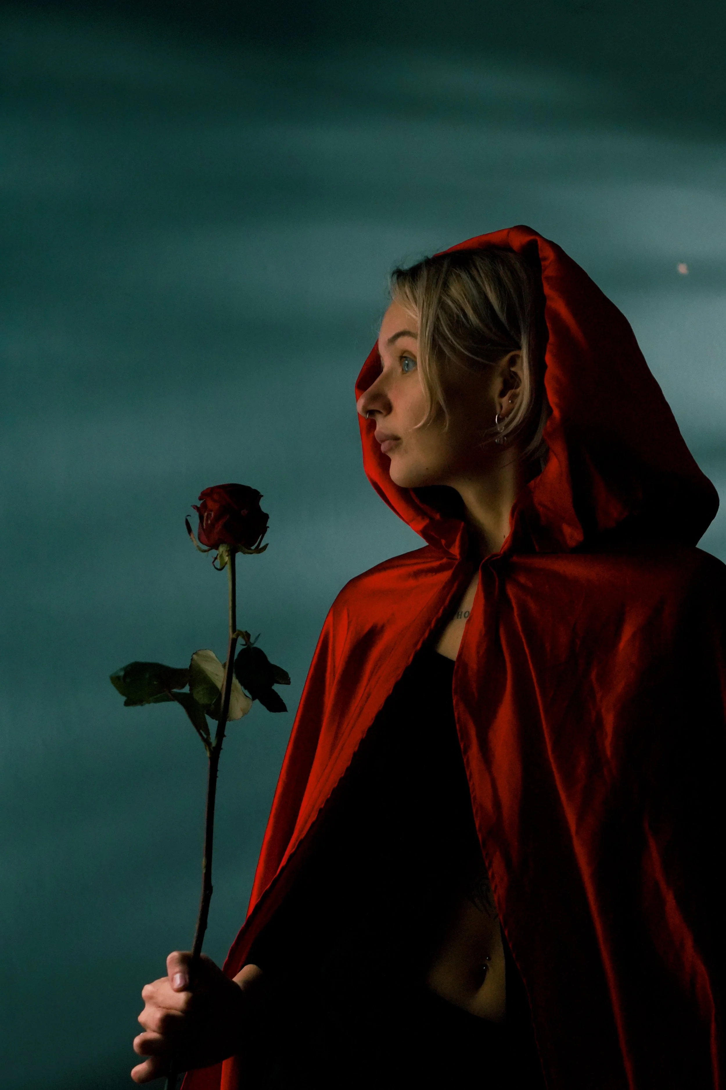 A woman with blonde hair wearing a red hooded cape holds a dark, withered rose against a dark, teal background.