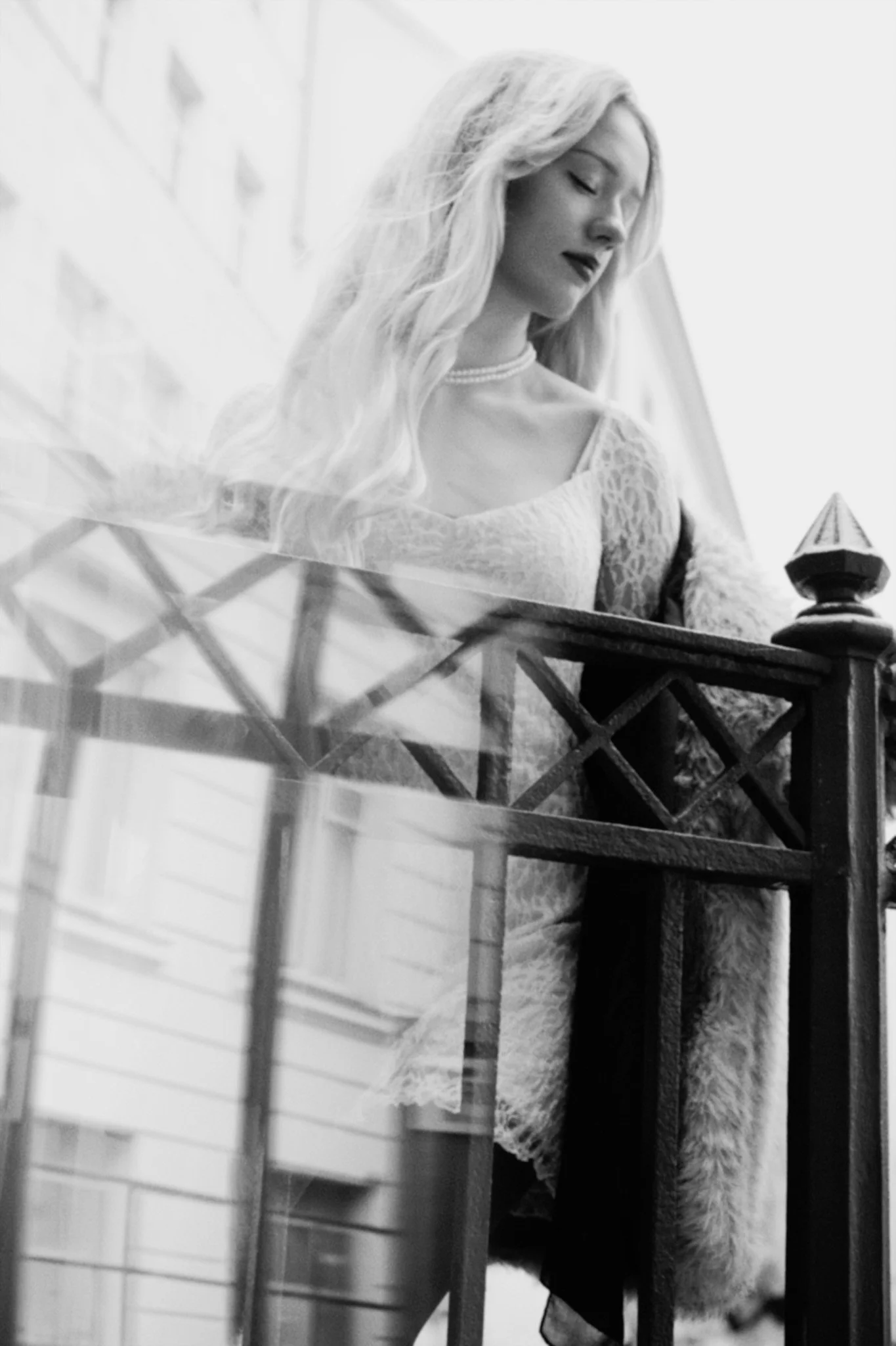 Eine Frau mit langen, welligen blonden Haaren und dunklem Lippenstift schaut nach unten, während sie auf einem Balkon oder einer Außentreppe an einem Geländer steht. Im Hintergrund sind städtische Gebäude zu sehen.