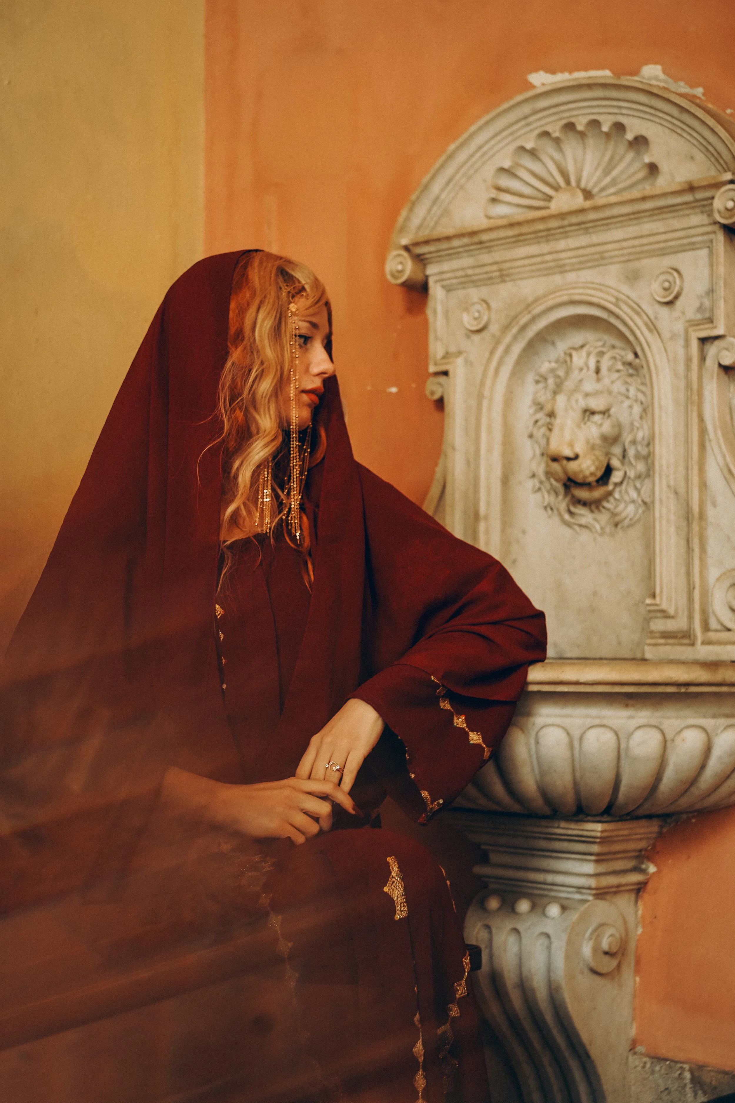A woman dressed in a red garment with gold embroidery, sitting beside a marble fountain with a lion's head carved on it, inside a room with orange walls.