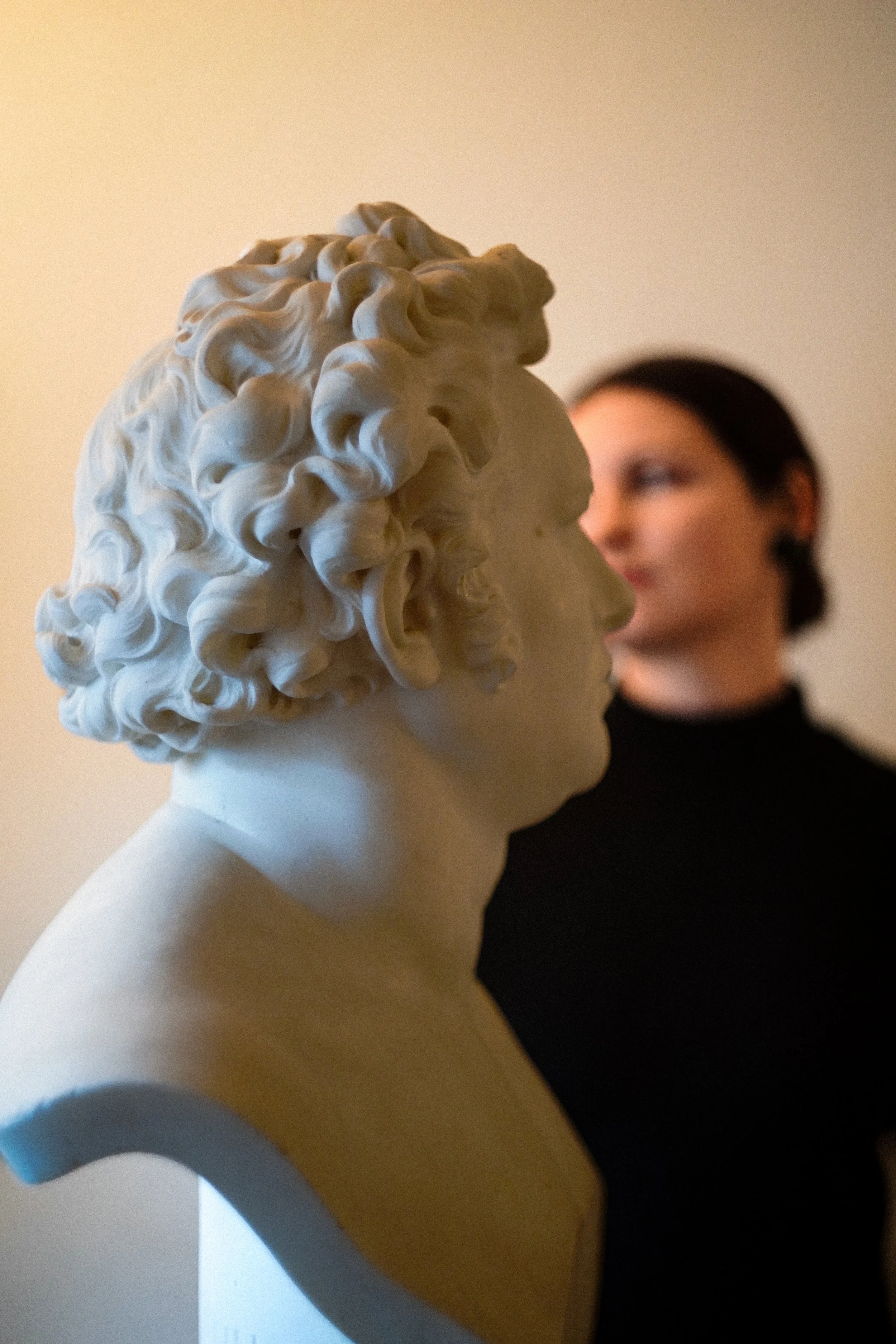 Close-up of a classical white marble bust of a man with curly hair, with a woman with dark hair and black shirt in the background out of focus.