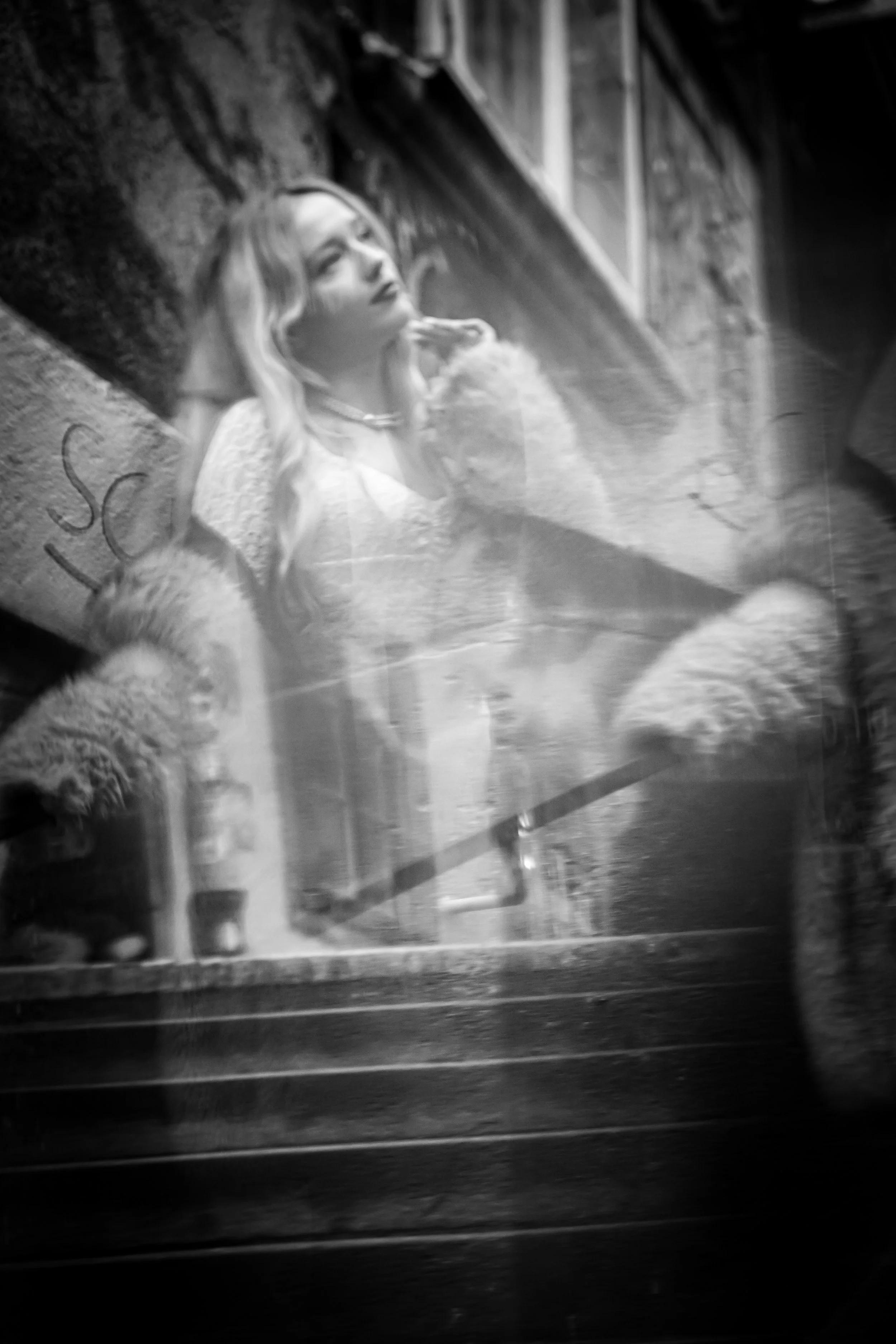 A woman with long hair and a fur coat sitting inside a cafe, seen through a window with reflections.