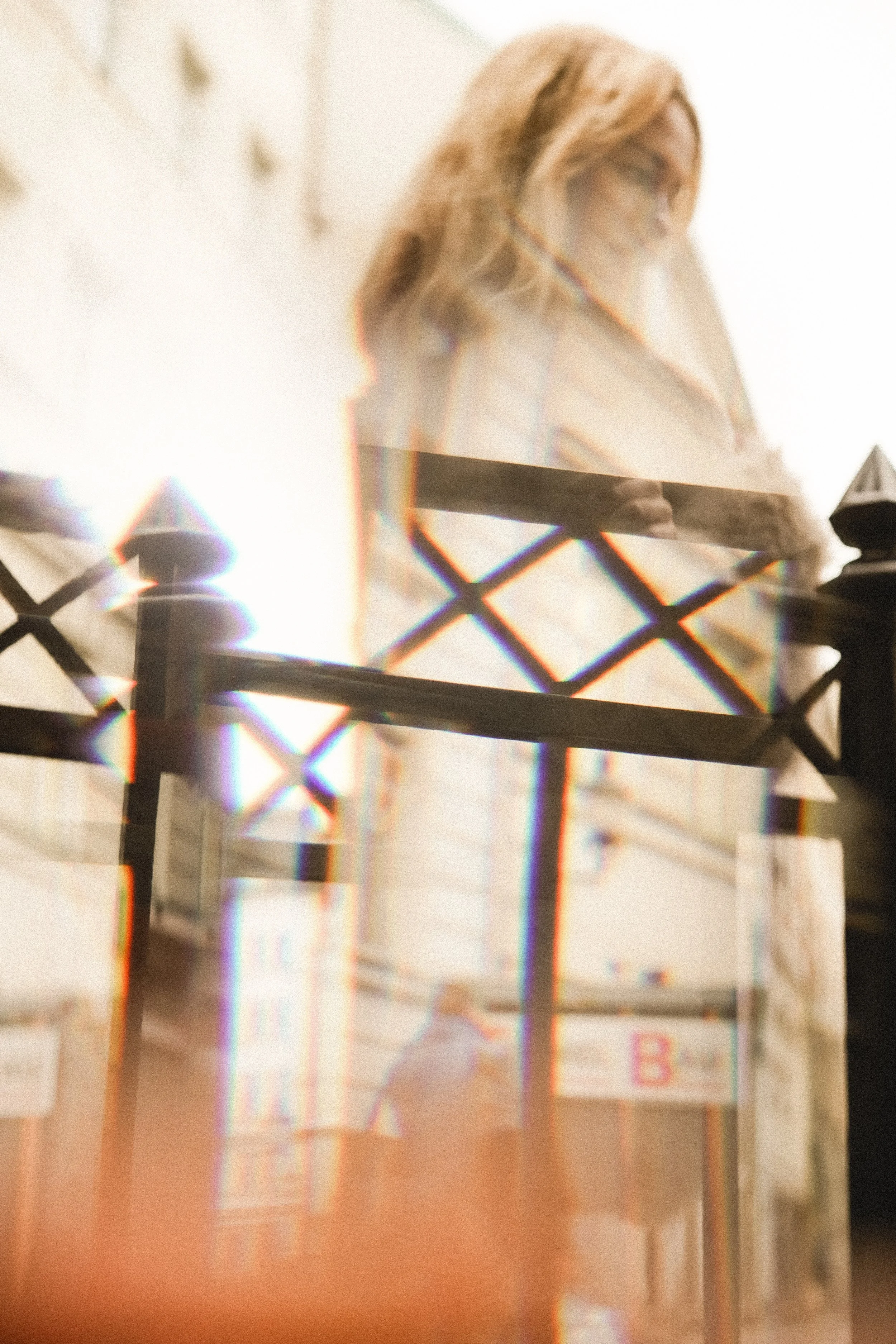 A woman with blonde hair and glasses looking down, seen through a decorative black fence with diamond shapes.