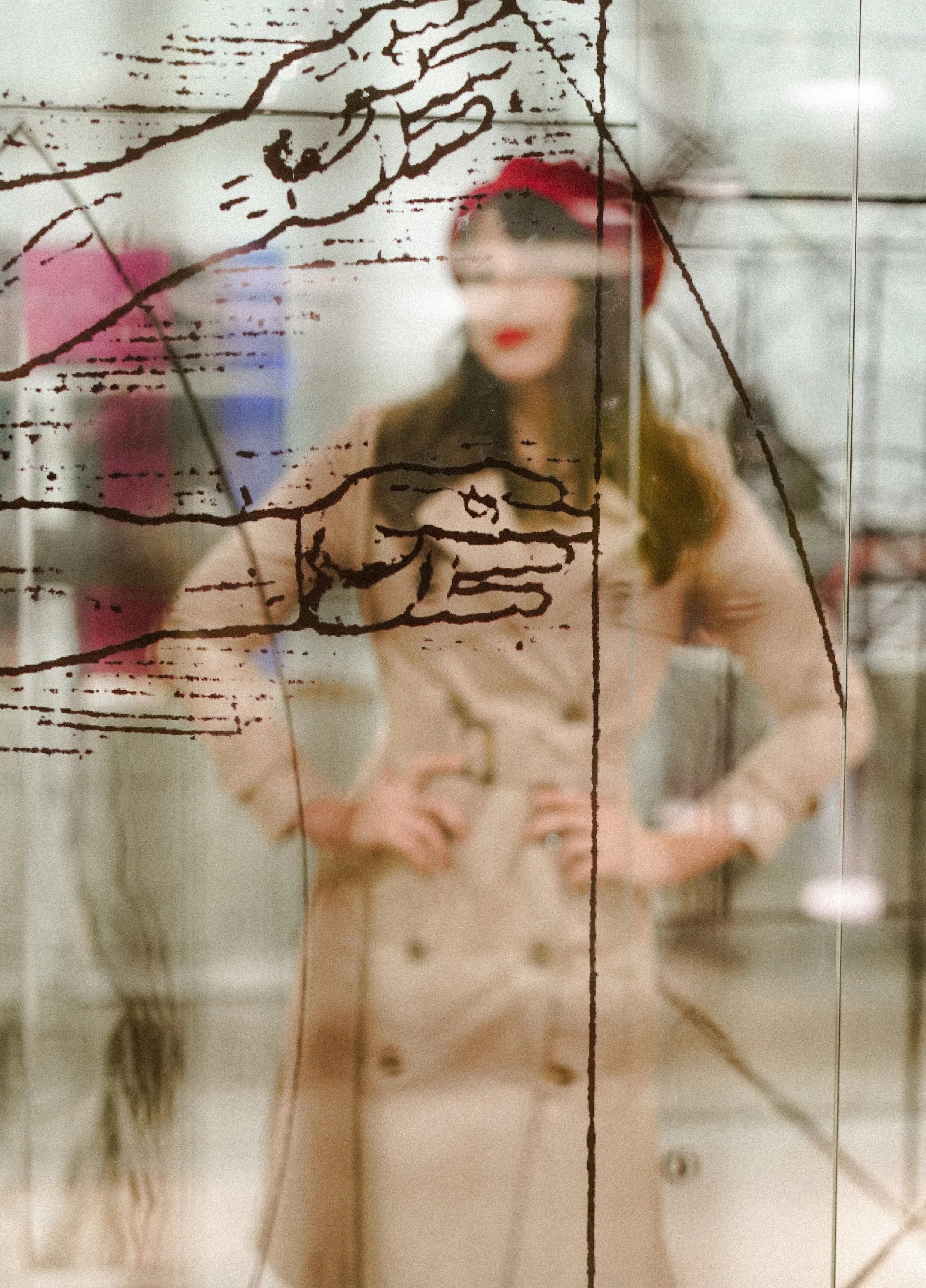 A woman with dark hair, wearing a red beret and a beige coat, stands with her hands on her hips behind a glass panel with a black marker drawing of a hand and part of an arm.