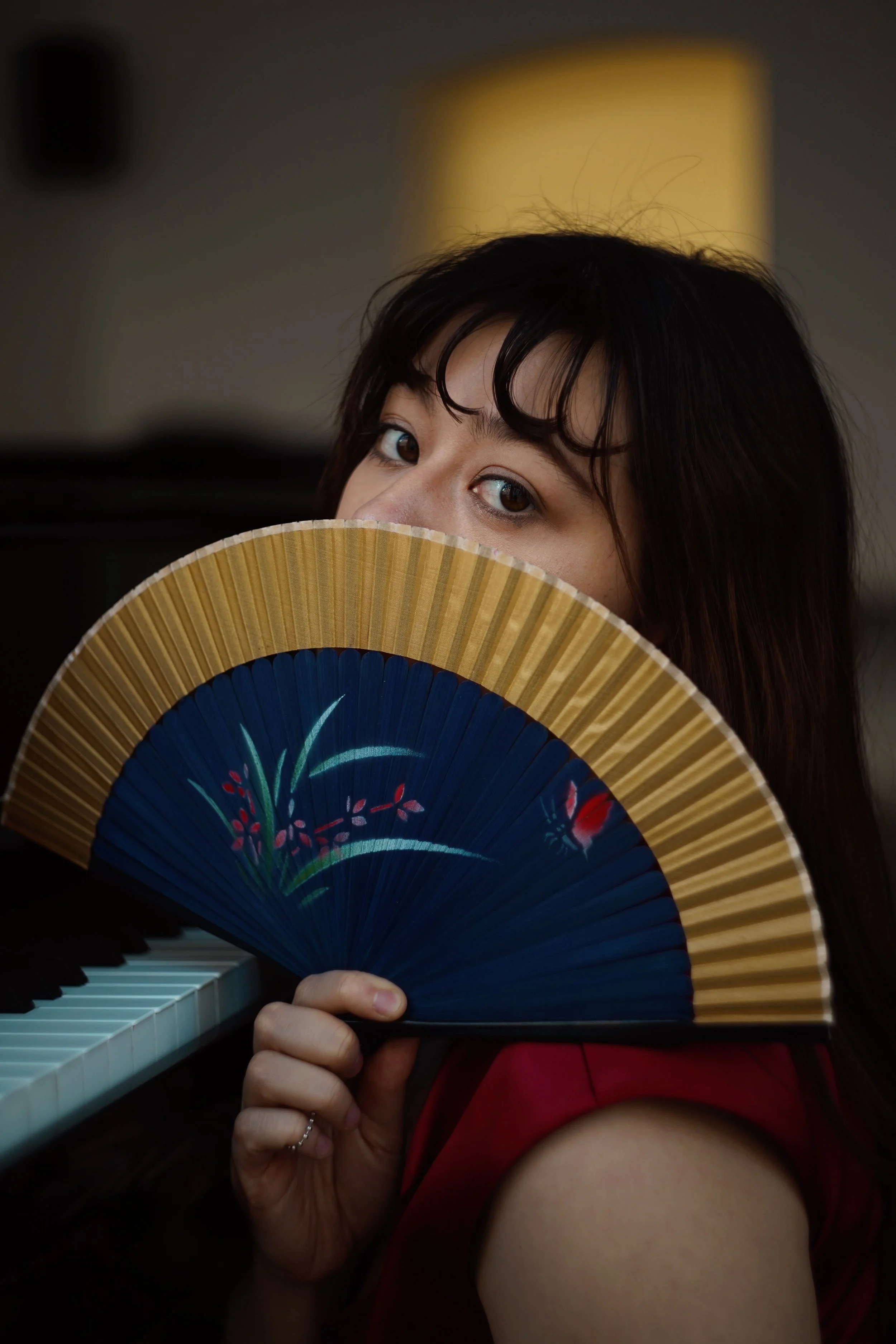 A woman with dark hair and eyes holding a decorative paper fan in front of her face, partially covering her mouth, with a piano and a yellow lampshade in the background.