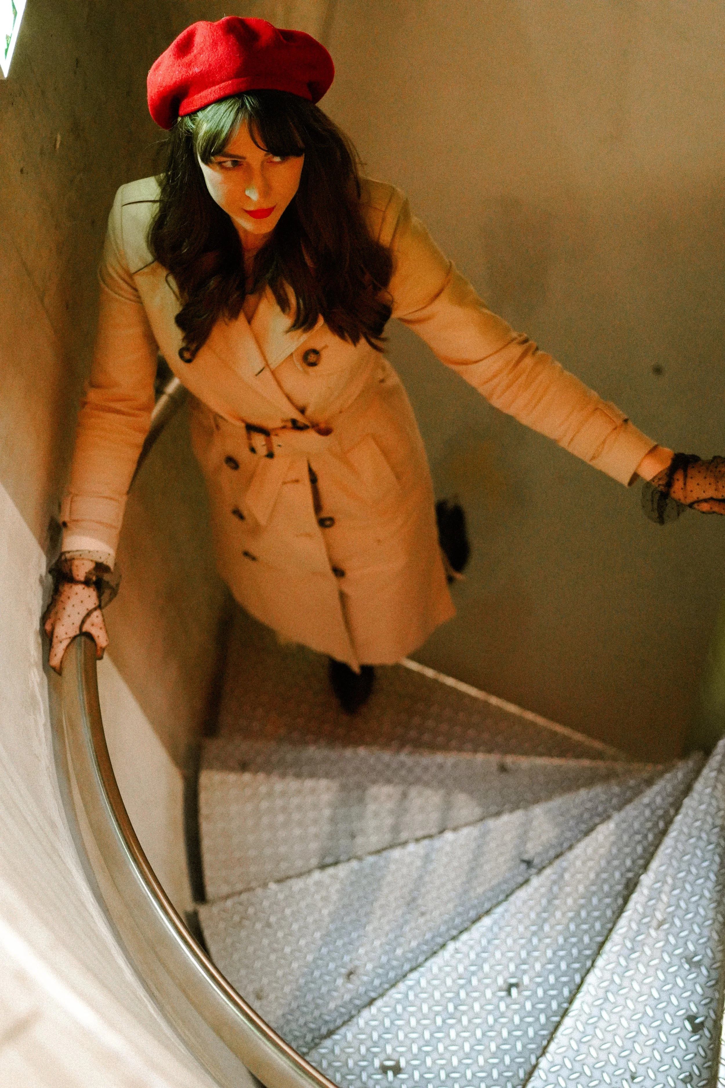 A woman with dark hair and bangs, wearing a red beret, a beige trench coat, and black polka dot gloves, standing on a spiral staircase, looking up towards the camera.