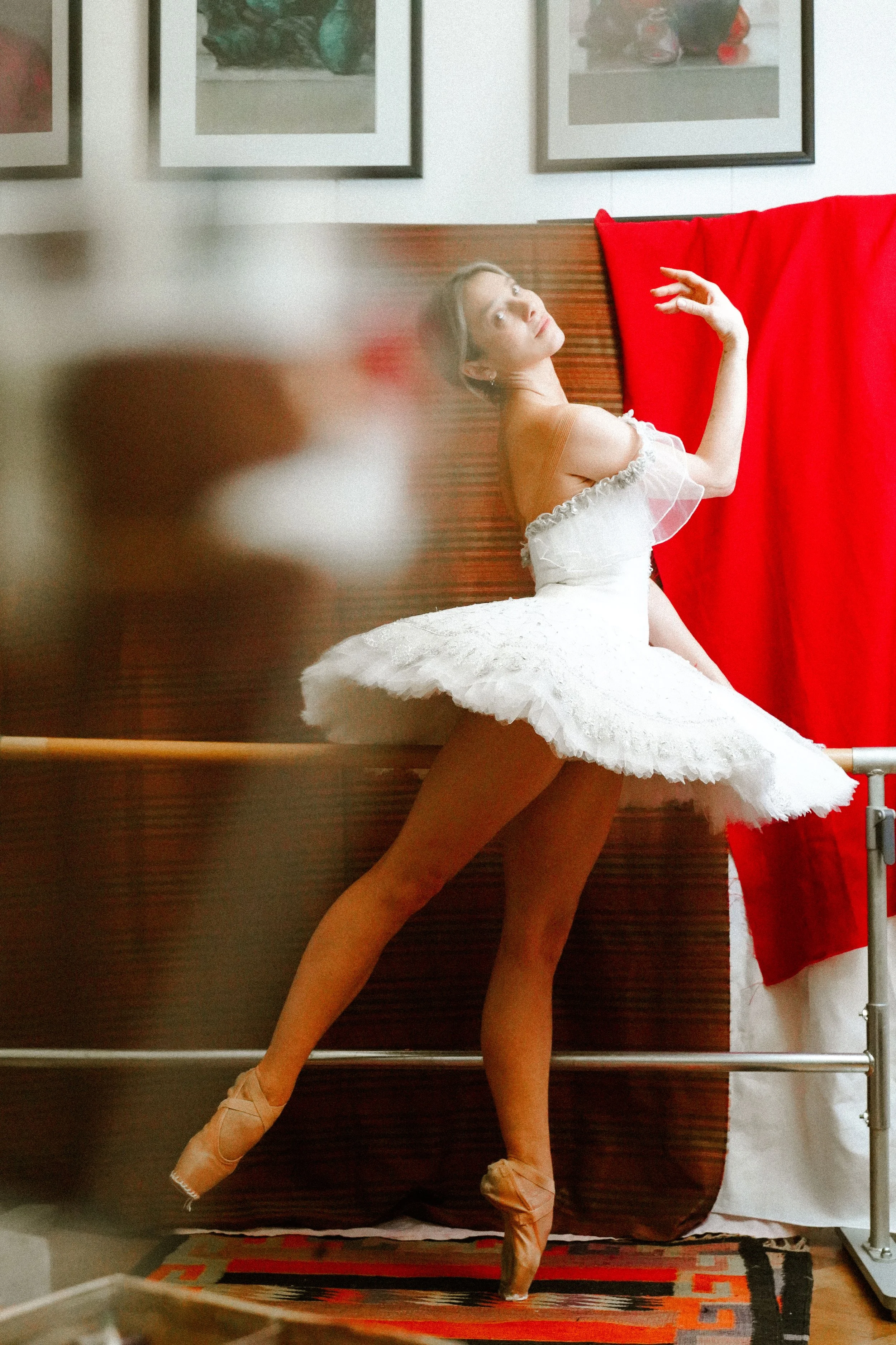 Ballet dancer in a white tutu and pointe shoes performing en pointe on one foot against a room with framed artwork and a red curtain.