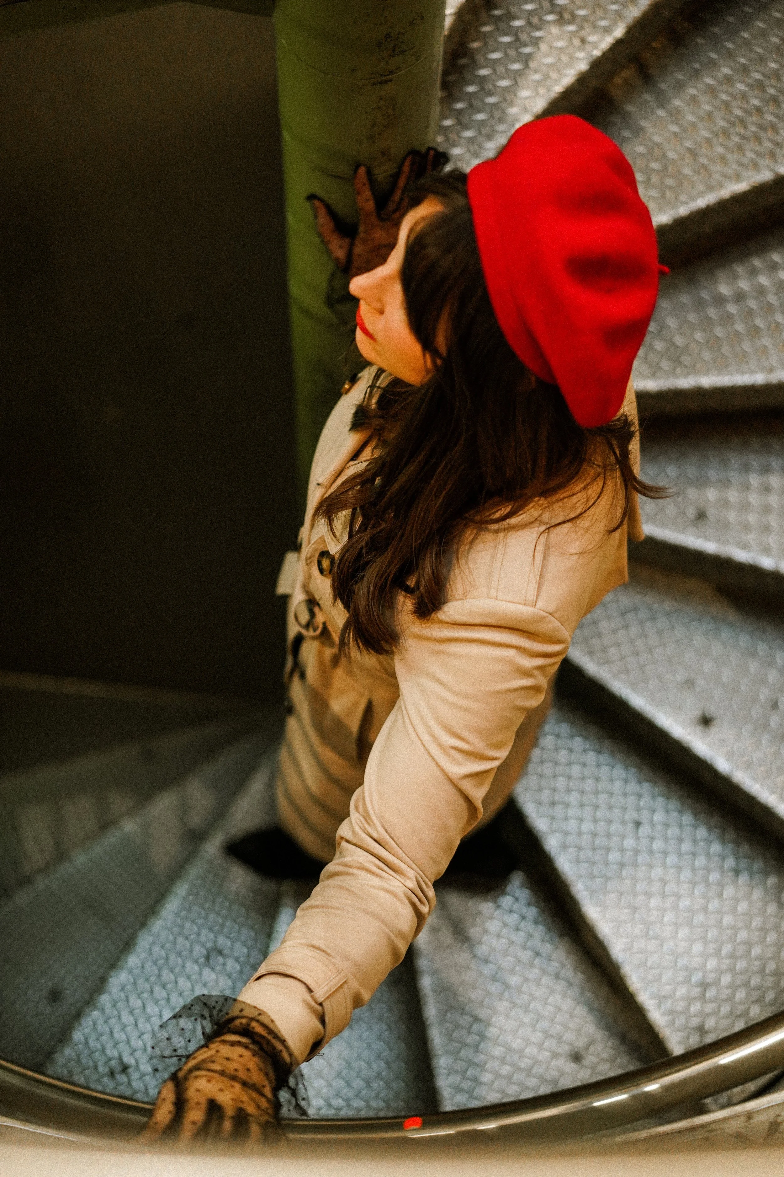 A woman with dark hair and a red beret sitting on metal stairs, looking up, wearing a beige coat and patterned gloves.