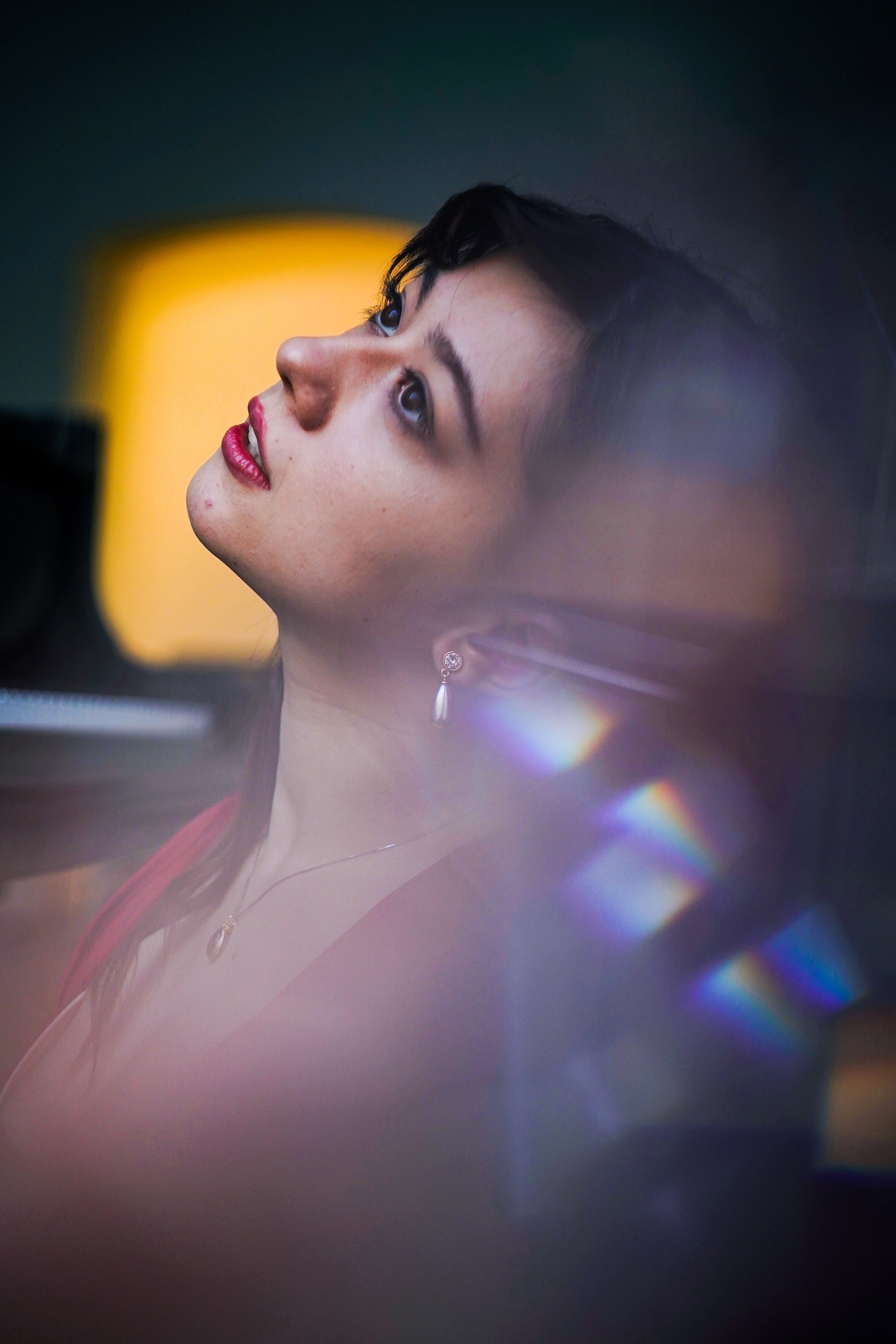 Close-up of a woman with dark hair, wearing earrings and a necklace, looking upward with a thoughtful expression and a blurred yellow light in the background.