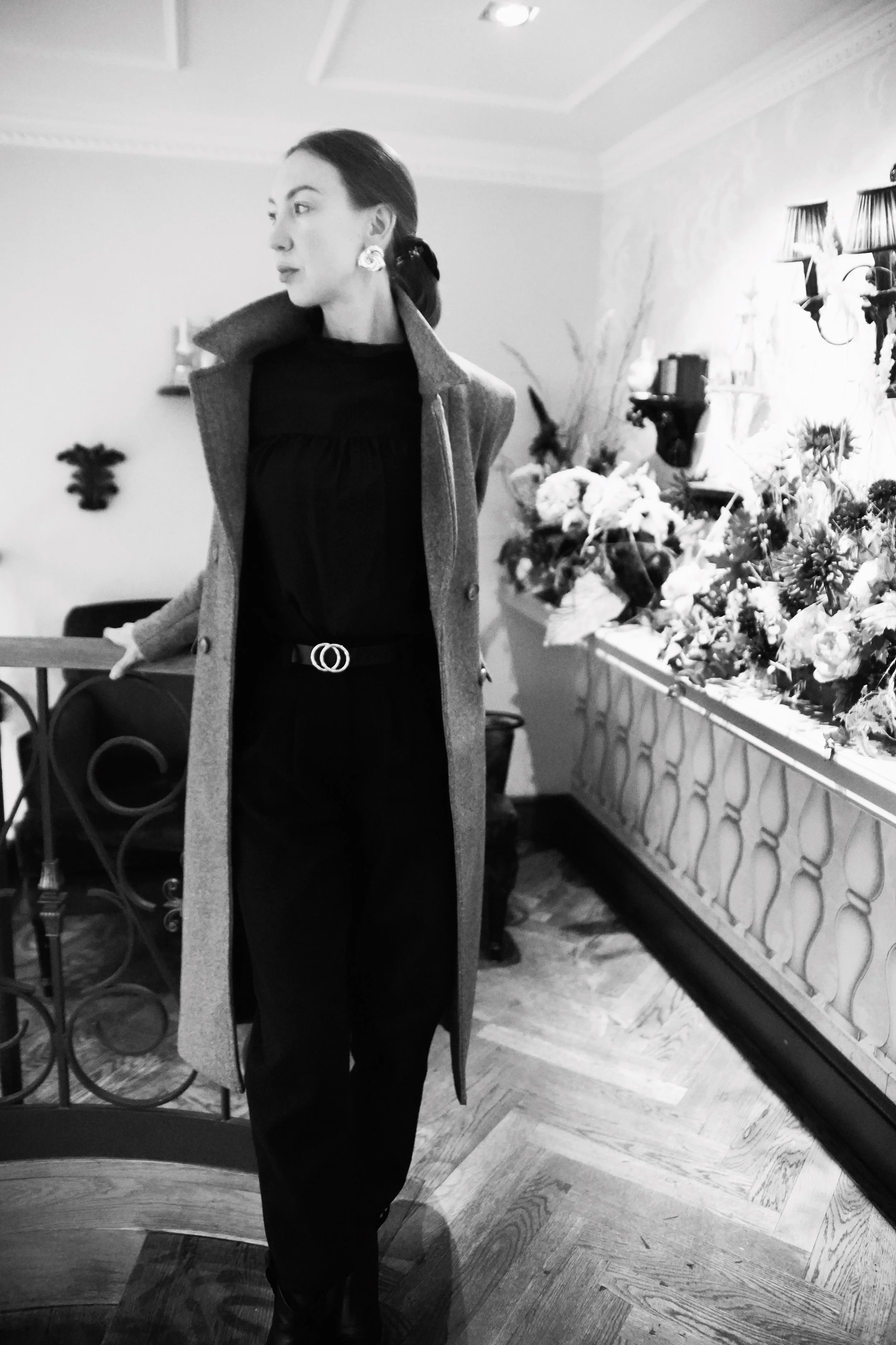 A woman wearing a long overcoat and black outfit posing indoors near a decorated wall with flowers in a vase.