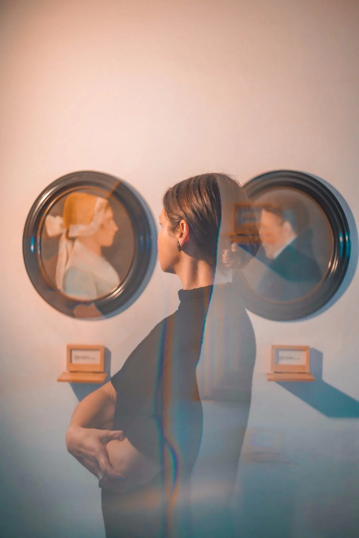 A woman with short dark hair wearing a black shirt and earrings is standing in front of two washing machines, looking at the reflection of a blonde woman with a bow in her hair in one of the machines.