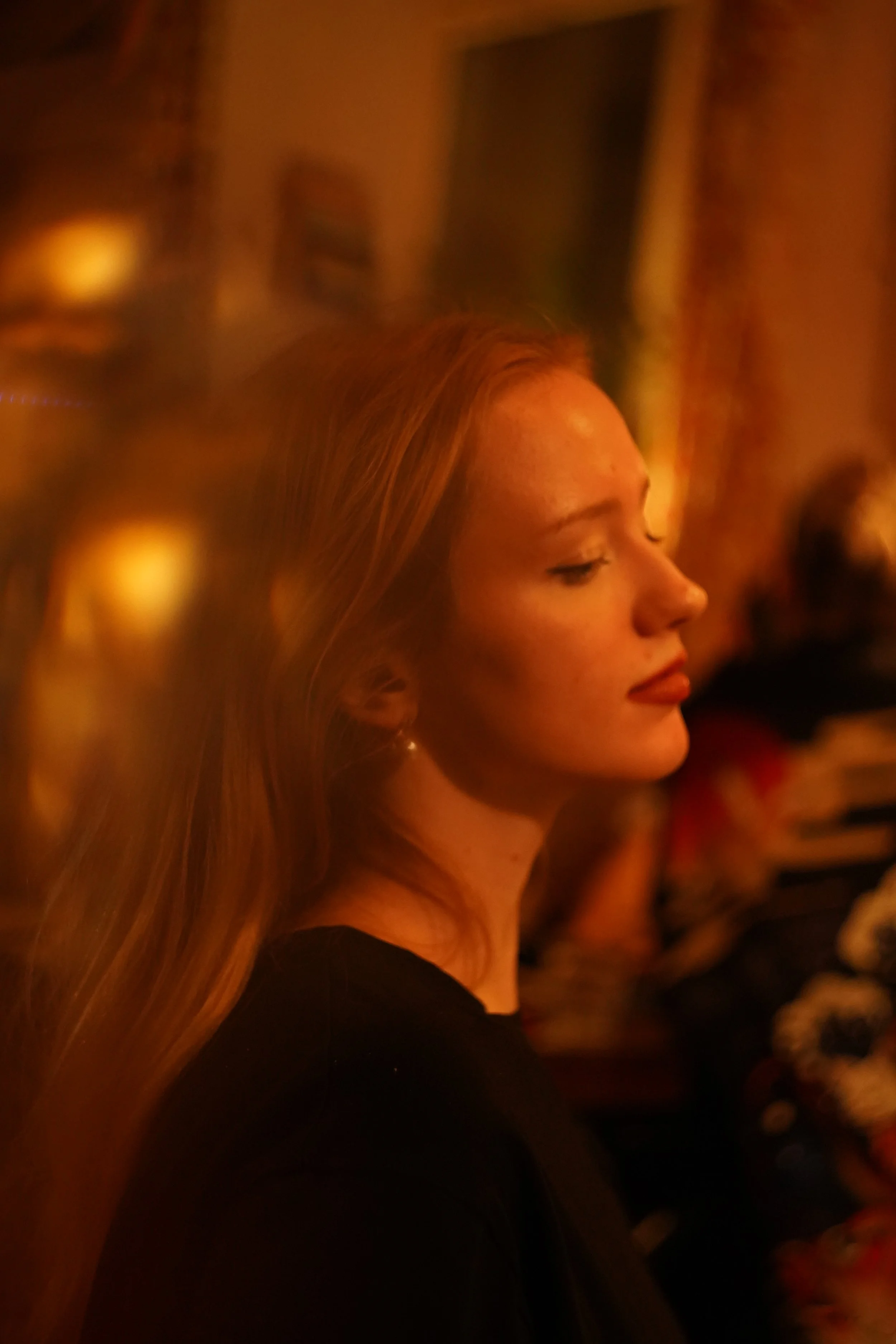 A woman with red hair and earrings is seen in profile, in a dimly lit, warm-toned room with a blurred background.