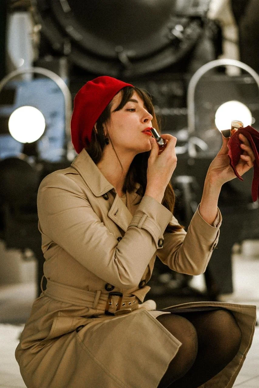A woman in a beige trench coat and red beret applying lipstick in front of a mirror in a dimly lit room.