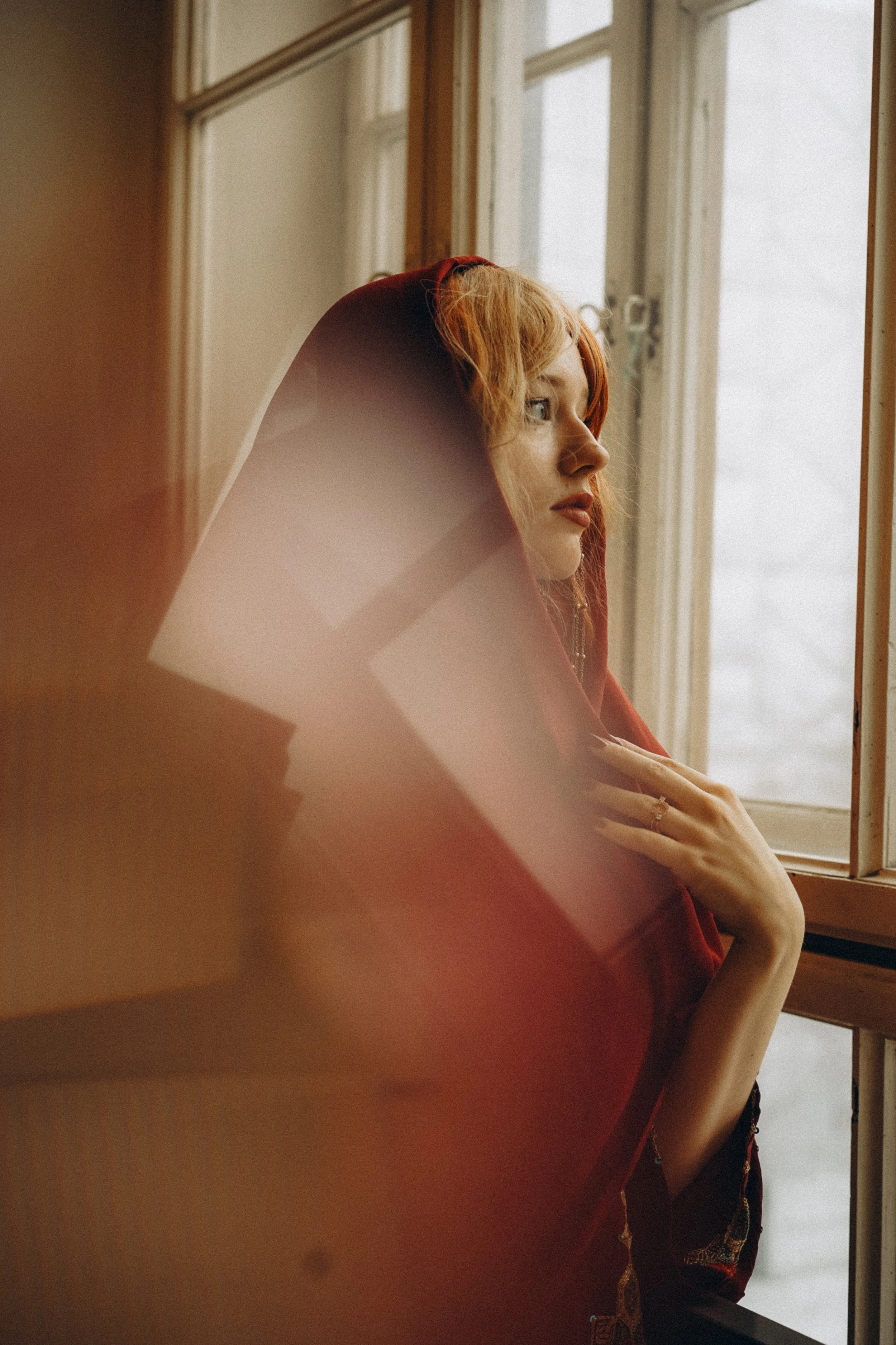 A woman with curly red hair gazes out a window, wearing a red hooded garment, with soft natural light illuminating her profile.