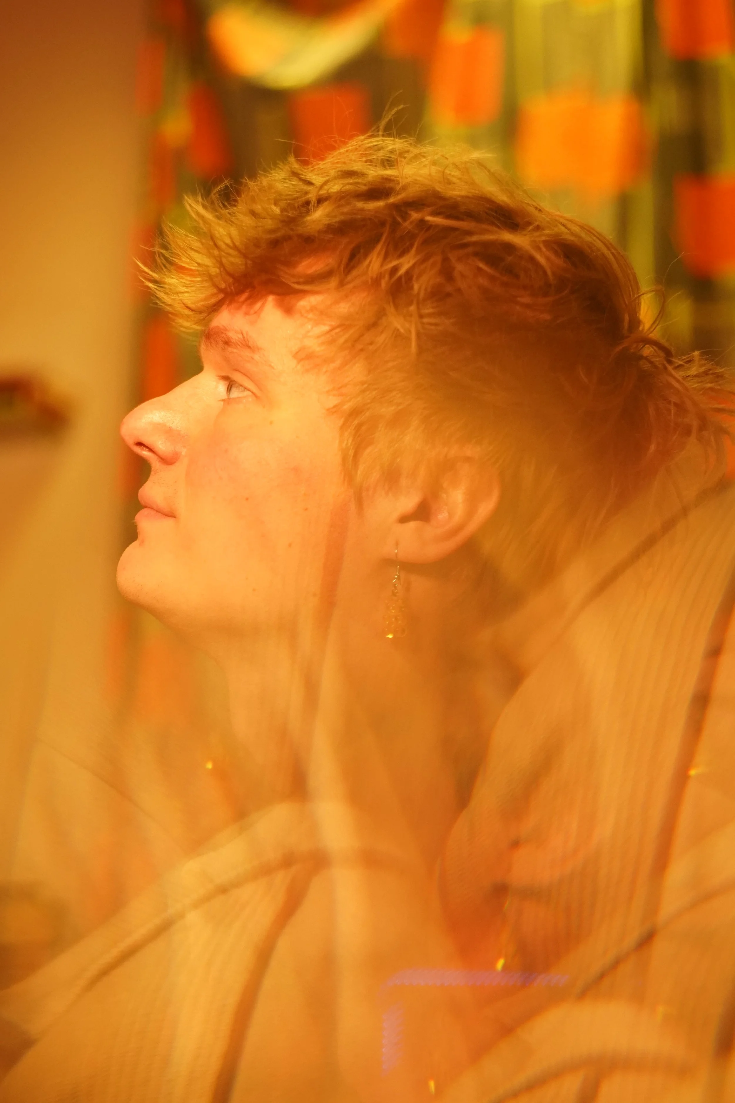 A person with short, curly red hair, sitting indoors with a background of colorful bookshelves, looking to the left.