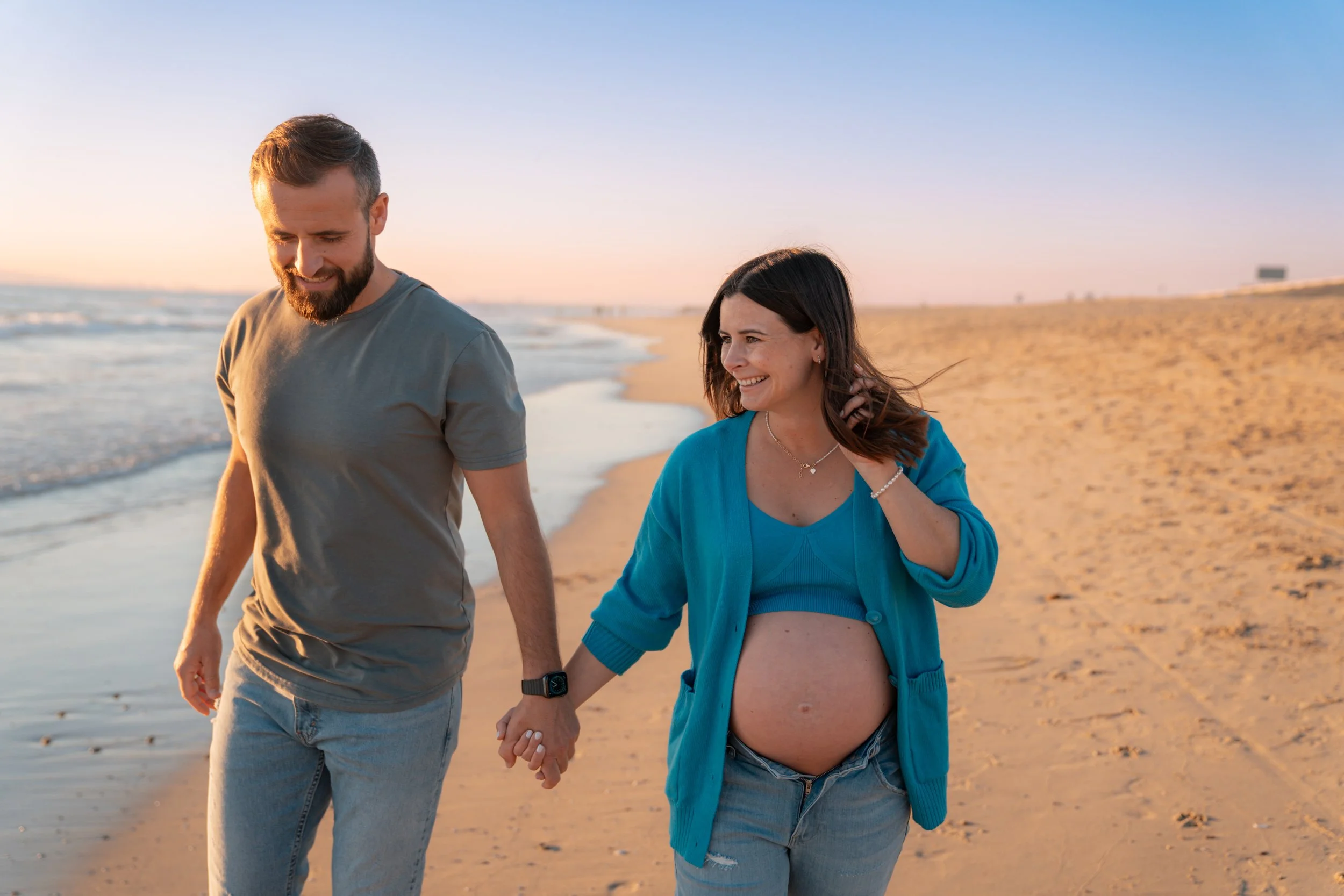 A pregnant woman and a man holding hands walk on the beach during sunset, smiling and happy.